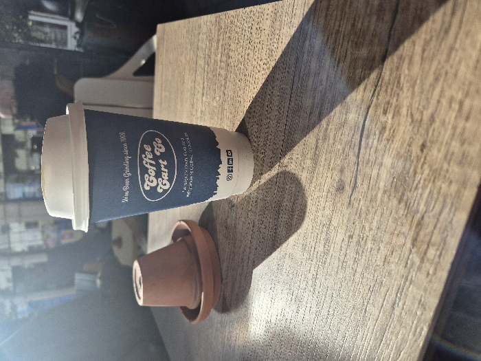 An indoor scene in Hereford, United Kingdom, displays a light brown wooden table surface. On the table, a blue disposable coffee cup with a white lid, prominently featuring "Coffee Cart Co" branding and the text "Hereford's Own Roasted Independent Coffee Roasters," stands upright. To the left of the cup, a small, inverted terracotta pot rests on a terracotta saucer. Direct sunlight from the left illuminates the foreground, creating distinct shadows of the cup and pot on the table. The background is out of focus, revealing blurred shapes consistent with an indoor public space, including indistinct figures and structural elements. No overt actions or interactions are visible.