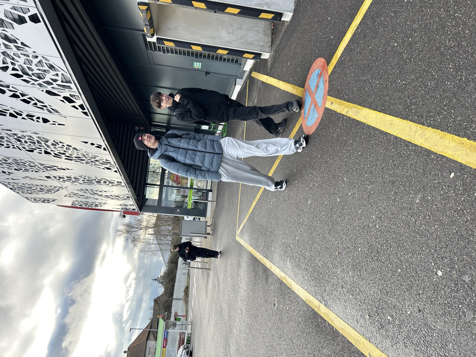 The scene depicts an outdoor paved area adjacent to a modern commercial building in Hœnheim, France, under an overcast sky. Three individuals are present. In the foreground, two young males walk towards the left. The male closer to the camera wears a black beanie, a grey-blue puffer jacket, light grey sweatpants, and black sneakers, carrying a dark backpack. He looks slightly to his right with a subtle smile. The male beside him wears a dark long-sleeved top, dark pants, and dark sneakers, also with a dark backpack; he looks slightly to his left with his mouth open and his left hand slightly raised. In the midground, a third individual, dressed in black, walks further away, looking down at a mobile phone held in their left hand.
The ground is dark asphalt pavement with bright yellow painted lines forming a directional cross in the foreground. A circular red and blue "No Parking" sign is painted on the asphalt, partially obscured by the feet of the two foreground males. To the right, a large, multi-story building features a distinctive metallic gray facade with intricate cut-out patterns and a red vertical stripe. The ground level includes glass automatic doors, through which a "FRAIS" sign is visible, suggesting a supermarket. Dark grey building sections with yellow and black striped safety barriers are also present. In the background, to the far left, a white car, green structures, and a house with a reddish-brown roof are visible. A line of leafless trees spans the distant horizon beneath a heavy, cloudy sky.