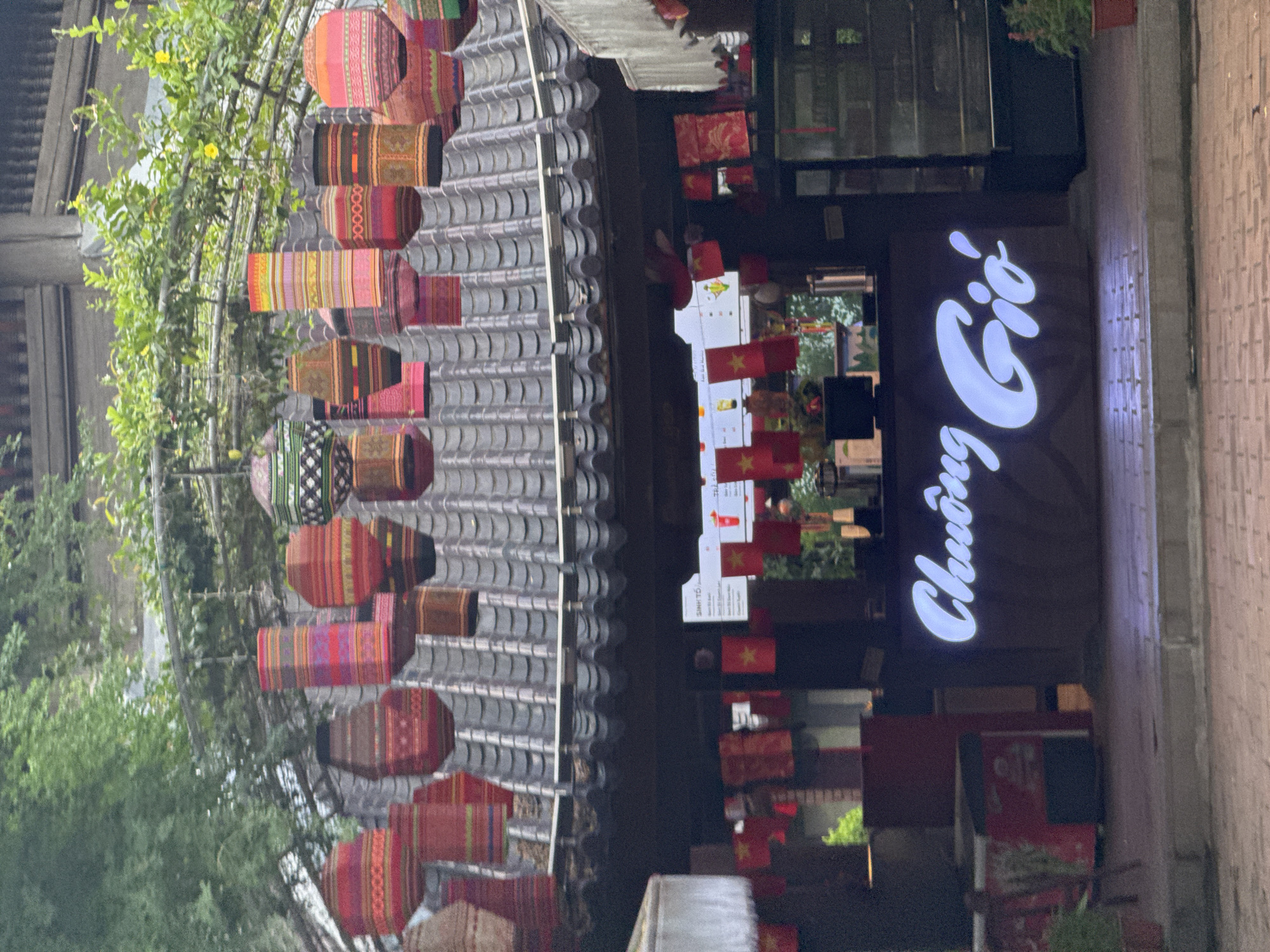 This is a daytime, eye-level, outdoor shot of what appears to be a market or cafe stall with a thatched roof and traditional Vietnamese lanterns hanging overhead. The lanterns are cylindrical and have intricate, colorful patterns. Below the roofline, small Vietnamese flags are strung together, indicating a potential patriotic or festive atmosphere. A counter with a glowing white sign that reads "Chuông Gió" is visible, suggesting the establishment's name. The flooring is paved with bricks. The environment is green, with trees and foliage visible in the background, hinting at a possible park or natural setting within the city of Tây Hoa Lư Ward, Vietnam.