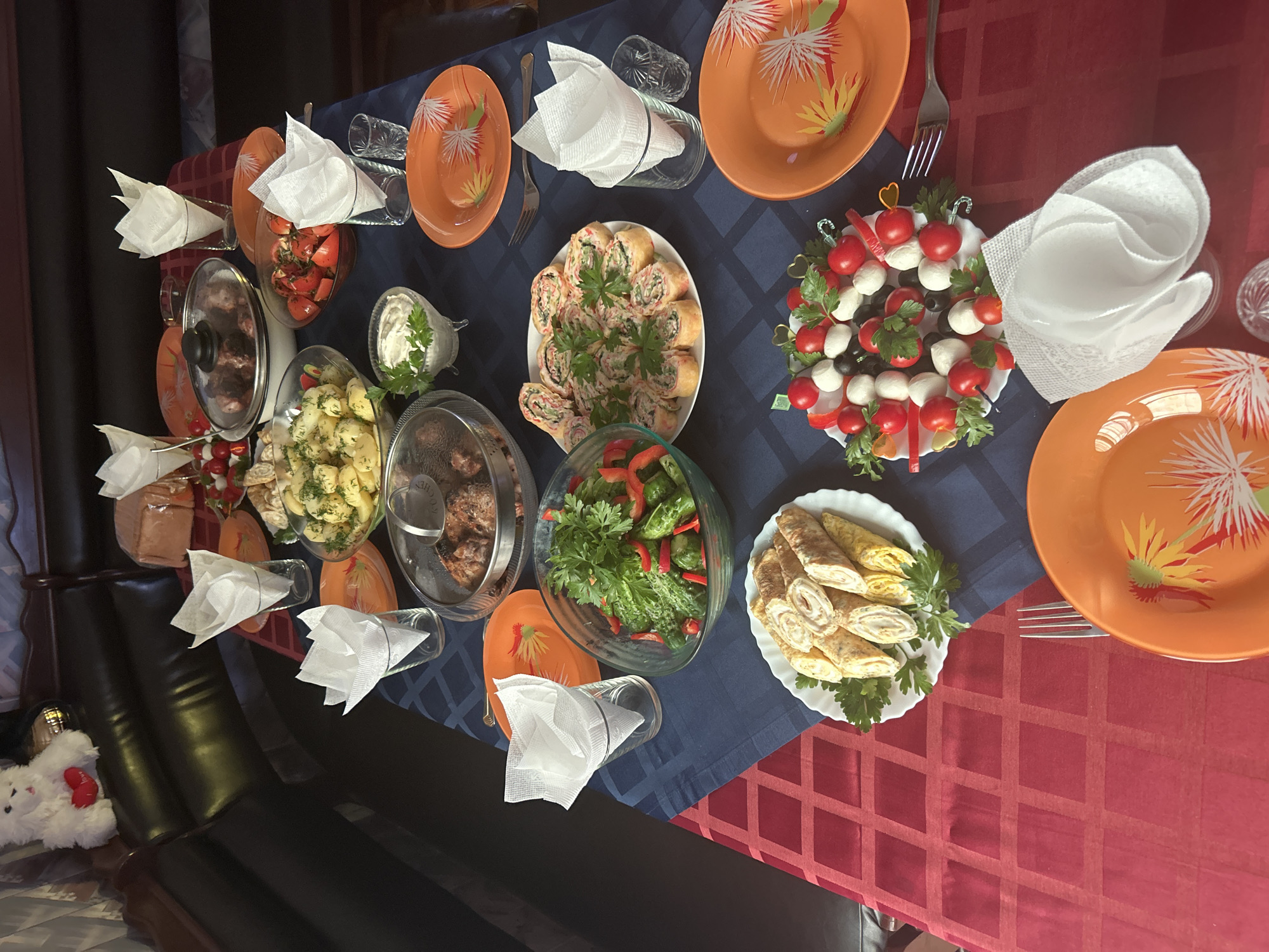 The image depicts a dining table set for a meal, likely a gathering or celebration, with a variety of dishes. The table is covered with a dark blue tablecloth with a geometric pattern and a red tablecloth on the left side. On the table are several plates of food, including what appears to be meatballs, a potato salad, a cucumber and pepper salad, sushi-like rolls, and small skewers with cherry tomatoes and mozzarella balls. There are also glasses with white napkins, orange plates, and cutlery. In the background, a white plush toy with a red heart is visible on a dark leather sofa. The overall impression is of a home-cooked meal being served. The location is specified as Gelendzhik, Russia, suggesting a domestic setting in that city. No people are visible in the image.