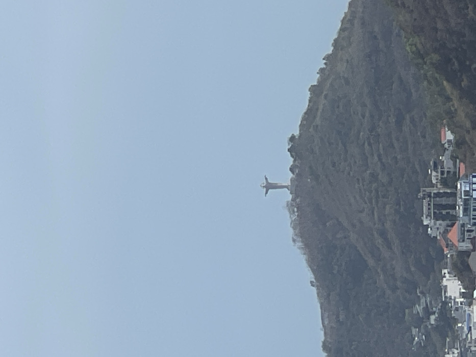 The image shows Christ the Redeemer statue atop Corcovado Mountain. A vast, light blue sky contrasts with the textured, verdant mountain. Buildings dot the lower right edge, suggesting an urban setting. The statue, a light gray, stands prominently against the mountainside, its arms outstretched.