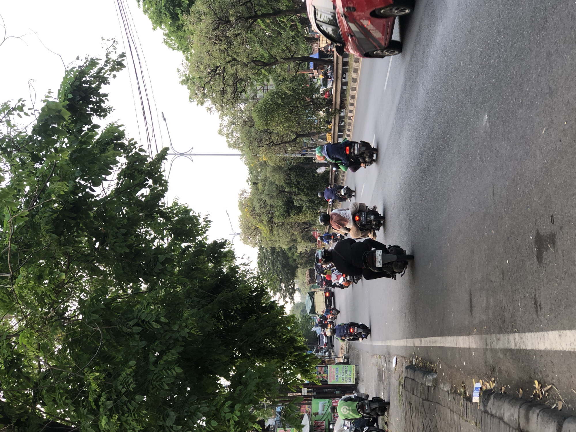 The image depicts a street scene in Kludan, Indonesia, characterized by moderate traffic flow. A red car is in the right foreground, moving away from the viewer. The road is paved and marked with white lines. Numerous motorcycles are visible, with several individuals riding them, some with passengers. Some riders are wearing helmets and casual clothing, while others have on darker attire. Trees with lush green foliage line the street on both sides, and utility poles with wires are present. Signs are also visible along the roadside, one of which advertises food items. The sky is overcast, suggesting a cloudy day. The overall impression is a typical urban street with active transportation.