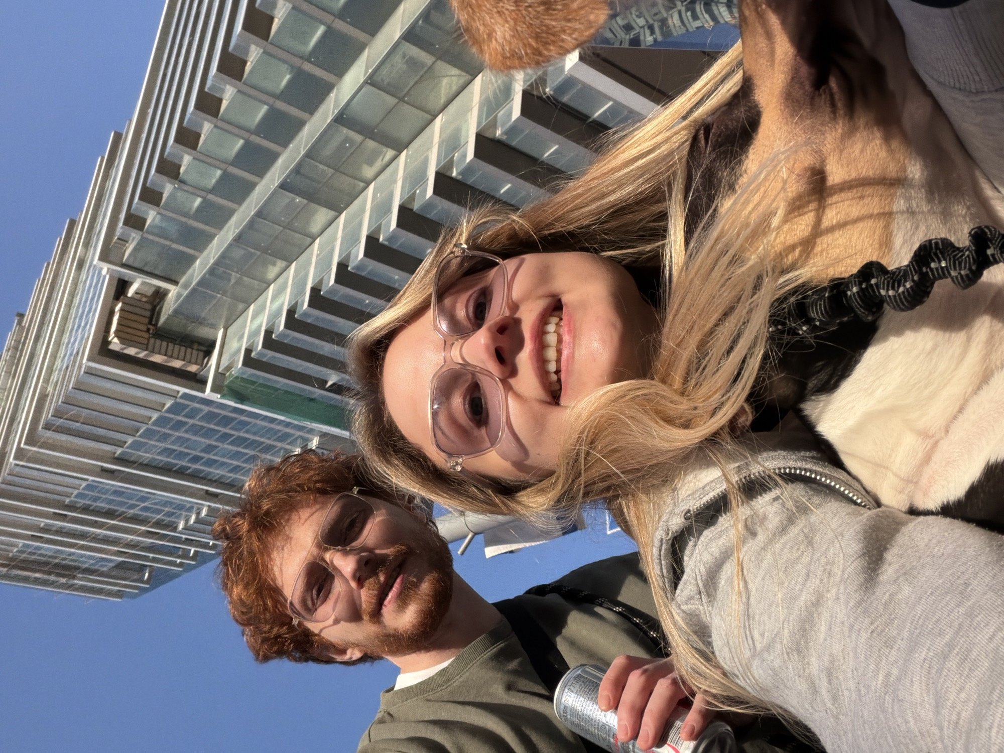 A low-angle selfie shows a woman and a man with a dog in front of a modern building. The woman, with blonde hair and glasses, is smiling. The man, with red hair and glasses, is holding a drink can. A dog is in the frame. The building has a modern design, with multiple horizontal layers and a glass facade. The sky is blue. The location is likely Tel Aviv, Israel, based on architectural style.