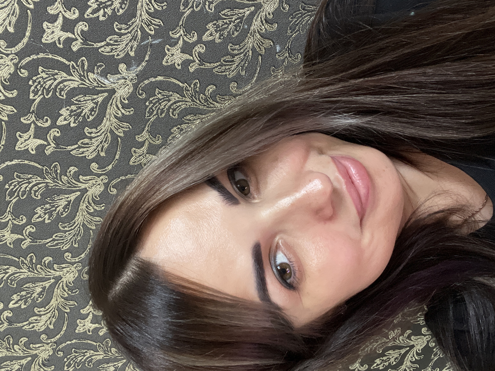 In a close-up, head-and-shoulders portrait, a woman with long, dark brown hair is positioned against a dark gray wallpaper featuring a gold floral damask pattern. The woman's face is turned slightly to her right, with her gaze directed towards the viewer. She has brown eyes, well-defined eyebrows, and her lips are slightly parted, revealing a subtle smile. Her skin appears smooth and lightly made up, with a hint of blush on her cheeks and a sheen on her lips. Her hair, which frames her face, has some lighter, possibly silver strands interspersed throughout. She is wearing a black top. The background wallpaper's repeating pattern is clearly visible, adding texture and visual interest to the scene.