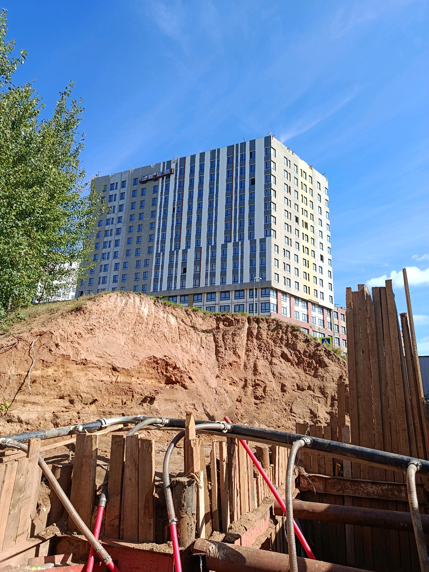 In Vnukovo, Russia, a multi-story apartment building under construction stands tall against a clear blue sky with scattered white clouds. The building features a modern facade with a pattern of alternating gray and white vertical panels, interspersed with numerous windows. A construction hoist is visible near the top of the building. In the foreground, a large excavated area reveals layered reddish-brown soil, indicating ongoing construction work. Wooden planks reinforce the sides of the excavation, and a network of black and red pipes snakes through the trench, suggesting utility work. To the left of the frame, a large green tree with dense foliage adds a natural element to the urban construction scene. To the right, a vertical wooden structure, possibly part of the construction site's temporary fencing or shoring, is visible.
