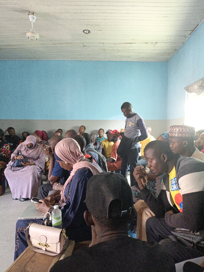 An indoor scene in Fagge C, Nigeria, depicts a gathering of approximately 20-30 individuals within a room featuring light blue walls above a grey wainscoting and a white paneled ceiling with a visible ceiling fan and a recessed light fixture. Natural light enters from a bright window on the right.
In the foreground, a person with their back to the camera, wearing a black baseball cap and t-shirt, is seated. To their right, another seated male wears a yellow t-shirt with a colorful graphic under a striped grey and black long-sleeved top; he holds a small, light-colored object in his right hand. A standing male, wearing a grey and blue long-sleeved shirt with a logo, is positioned slightly right of center, looking downwards.
Seated in the mid-ground, a woman wearing a light pink hijab and dark blue dress is focused on a white laptop and smartphone resting on a wooden surface in front of her. A clear plastic water bottle and a light pink handbag with a gold clasp are also on this surface. To her left, another woman in a light purple hijab and full-length dress, with a black face covering, looks down at an object in her lap.
Additional men and women are seated in rows behind these figures, extending into the background, some wearing traditional head coverings. The overall scene suggests a community meeting or gathering.