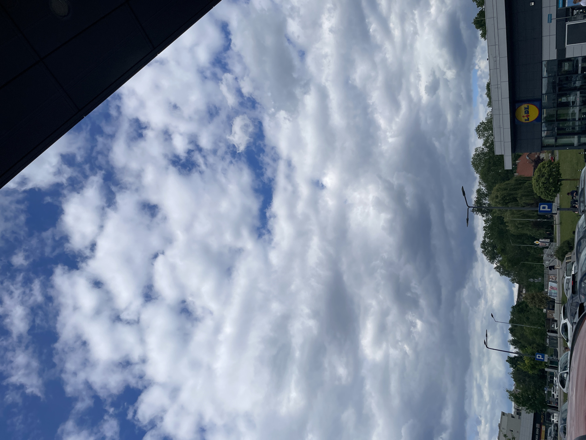 Here's a description of the image:
The image shows a sky filled with a mix of blue patches and predominant white and grey clouds. In the foreground, a dark, angled structure is visible. On the right side, there's a building with a "LIDL" sign, trees, street lamps, parked cars, and signs indicating a parking area. The location appears to be an urban environment.