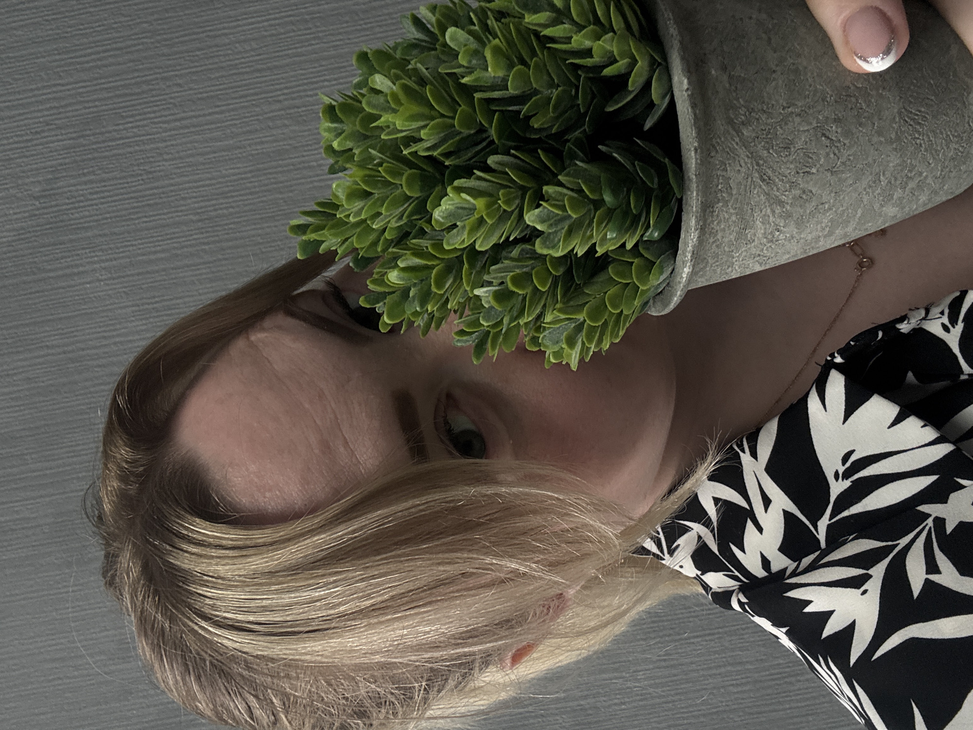 A fair-skinned woman with blond hair holds a potted green plant near her face. The pot is gray and textured. She wears a black and white patterned top and a thin gold necklace. The background is a light gray, textured surface. The location cannot be determined from the image.