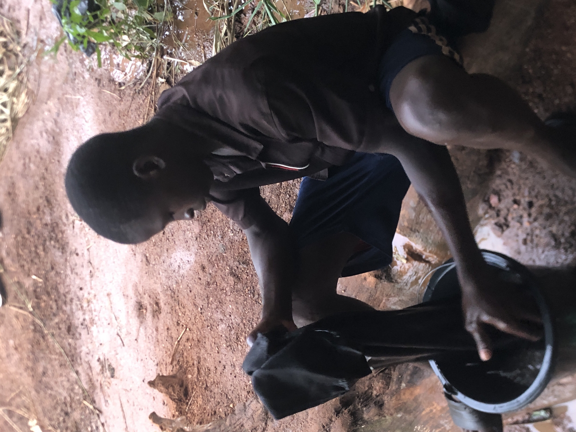 An individual, likely a young man, is seen outdoors in Ukwu-Abwa, Nigeria. He is kneeling beside a black bucket and appears to be rinsing or cleaning a dark-colored cloth within it. The ground is earthen, and there's some vegetation and water visible in the background. The individual is wearing a dark-colored shirt and dark shorts.