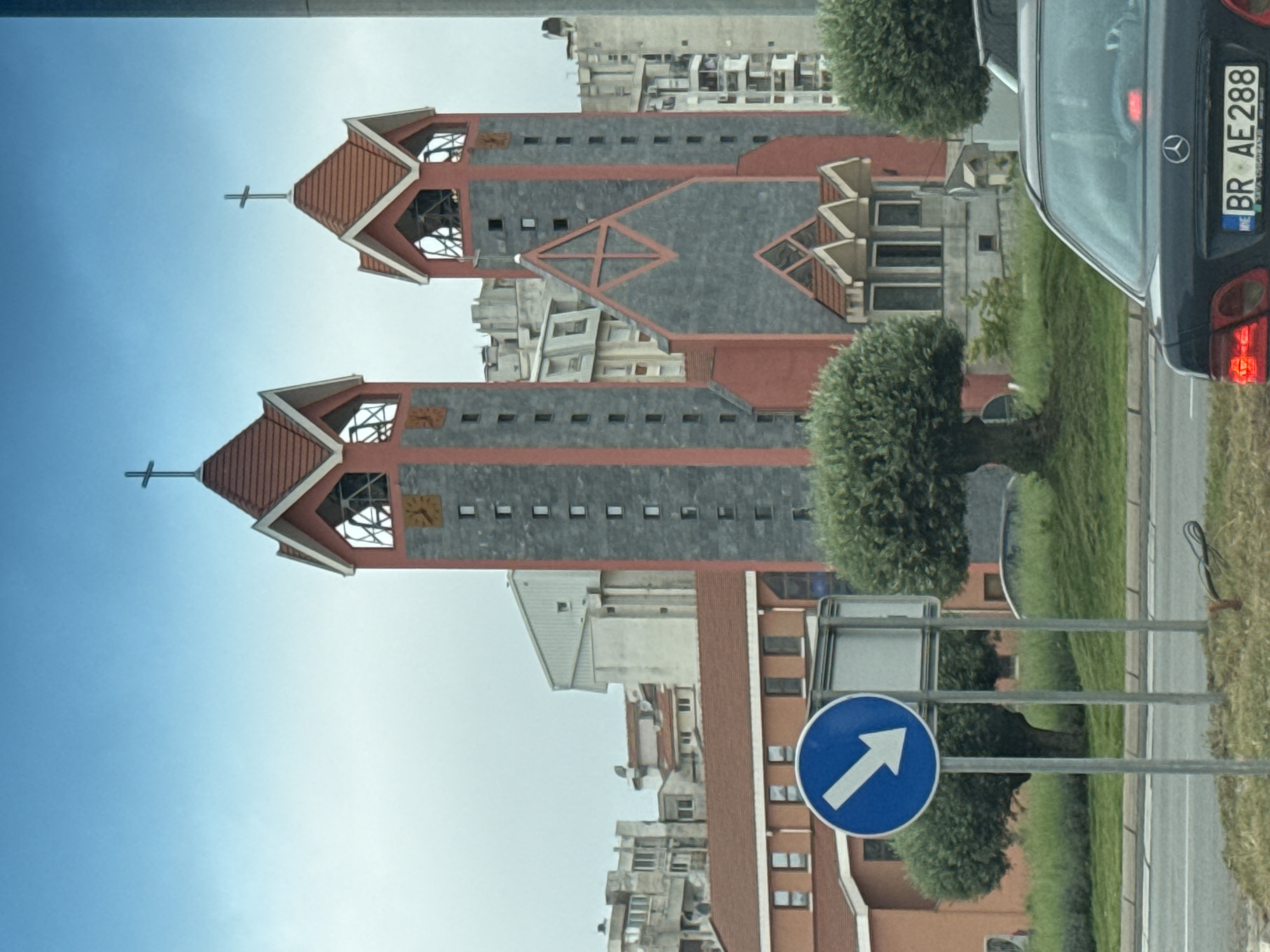 The image depicts the Church of St. Petka in Bar, Montenegro, from a street-level perspective. The church features two tall bell towers with terracotta-colored pyramidal roofs and crosses atop each. The towers have small windows and exposed framework at the top. A gray roof with red trim connects the towers, with a triangular design element in the middle. The church is surrounded by residential buildings. In the foreground, a blue sign with a white arrow pointing right is visible, along with a metal signpost. A black Mercedes-Benz car is also partially visible on the right side of the frame, with a license plate reading "BR AE 288". The sky is blue with light clouds. Grass and trees line the side of the road.