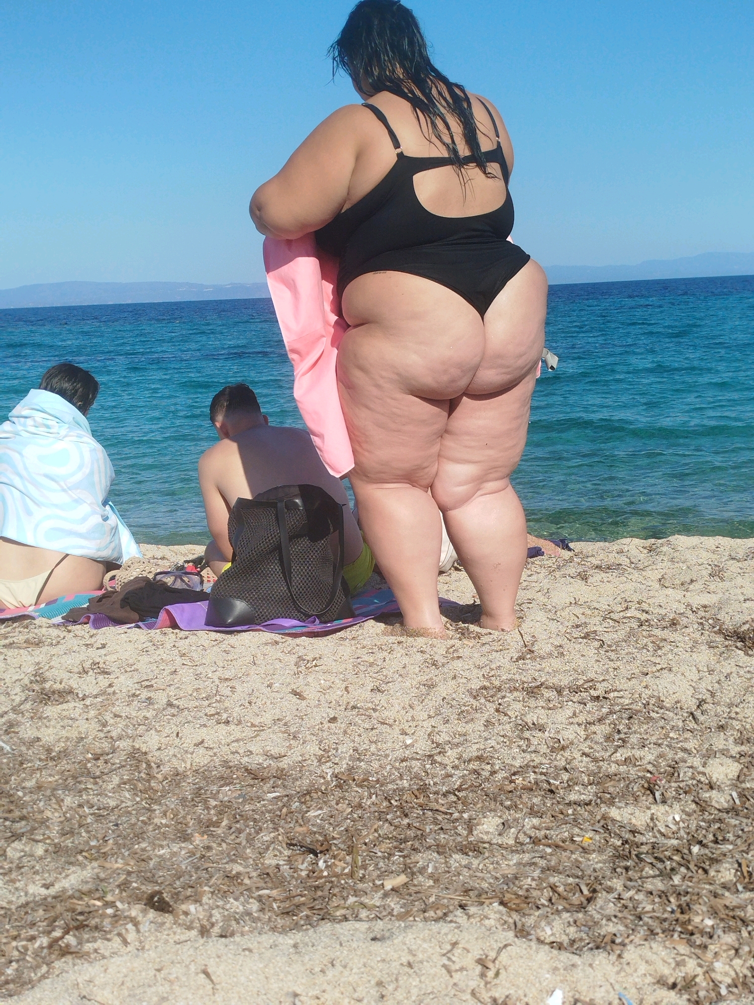 This image depicts a scene on a sandy beach in Sithonia Municipal Unit, Greece. In the foreground, a woman in a black swimsuit is holding a pink towel and facing away from the camera. Her back and legs are visible. Further back on the beach, two individuals are seated, one wrapped in a blue and white patterned towel and the other shirtless. A black mesh tote bag, a pair of goggles, and towels are spread out on the sand in front of them. The turquoise sea and a clear blue sky dominate the background. The shoreline is visible with some dried seaweed on the sand.