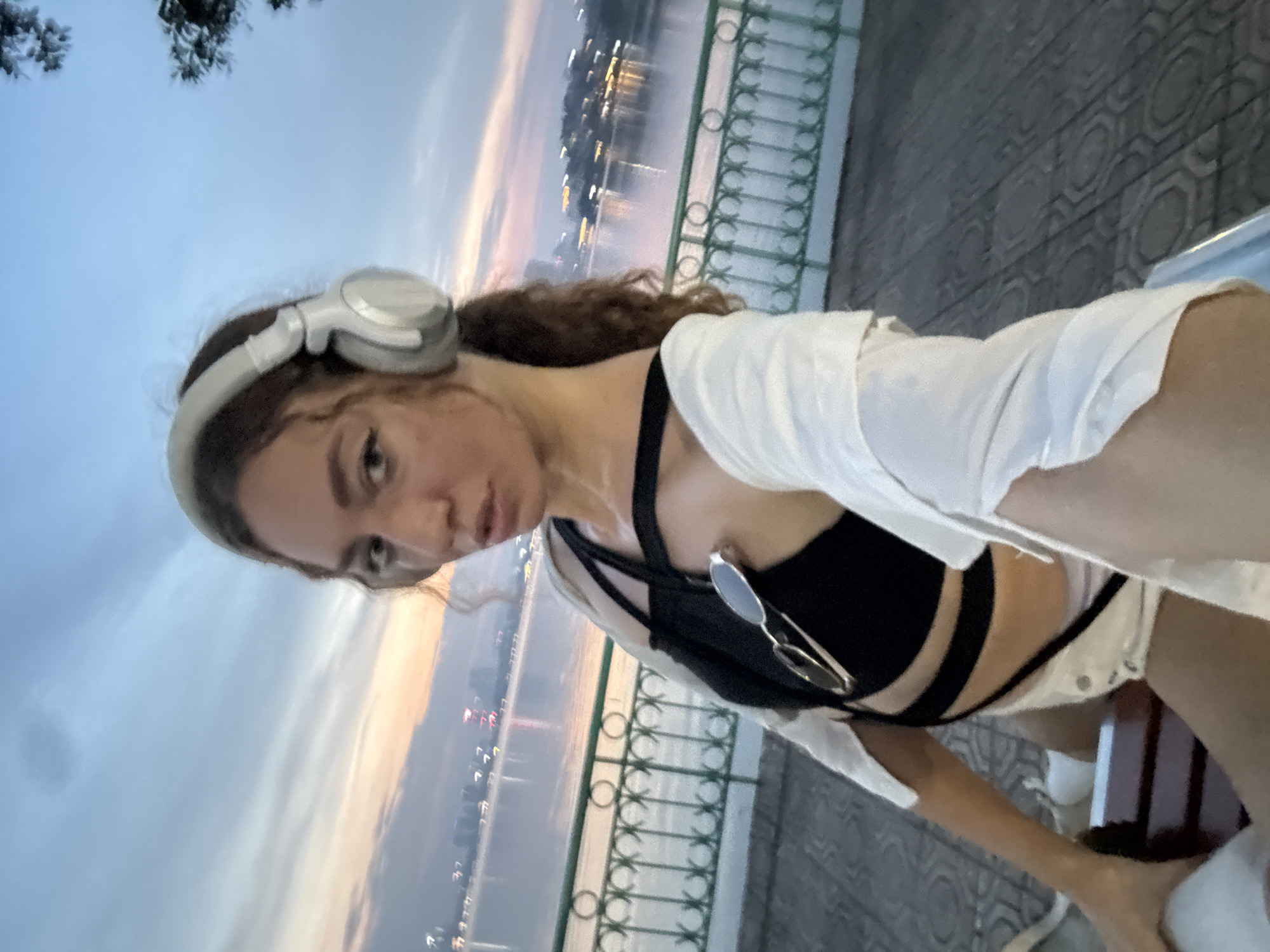 A young woman with curly brown hair, wearing white headphones and a black bikini top with a white shirt draped over her shoulders, takes a selfie. She is outdoors near a body of water, likely a river or lake, with a city skyline visible in the distance. The sky is overcast with hints of pink and orange, suggesting either sunrise or sunset. She is seated on a bench with a decorative metal railing behind her and a patterned paved walkway. Sunglasses are clipped to her bikini top. The overall impression is a casual, possibly post-activity moment, with the woman appearing relaxed and enjoying the view. The setting seems to be a public area with a scenic backdrop, typical of waterfront promenades.