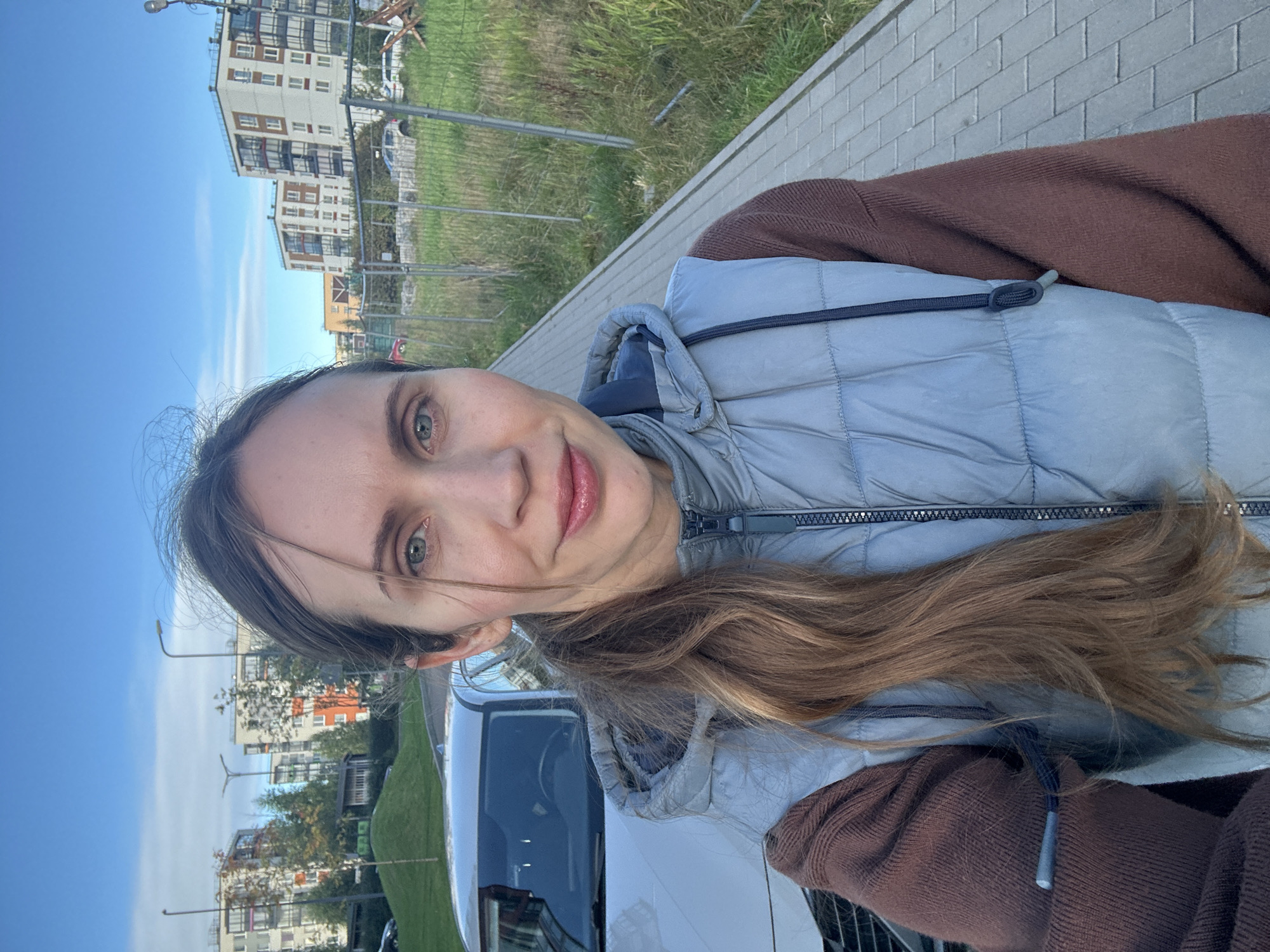 A woman takes a selfie outdoors. She is wearing a brown long-sleeved shirt and a light blue puffer vest. Her light brown hair is pulled back into a ponytail. She is standing on a paved walkway next to a silver car. In the background, there are apartment buildings, streetlights, and a grassy area with a chain-link fence. The sky is blue with some clouds. The location appears to be an urban residential area in Vsevolozhsk, Russia, possibly during autumn given the foliage and the type of clothing worn.