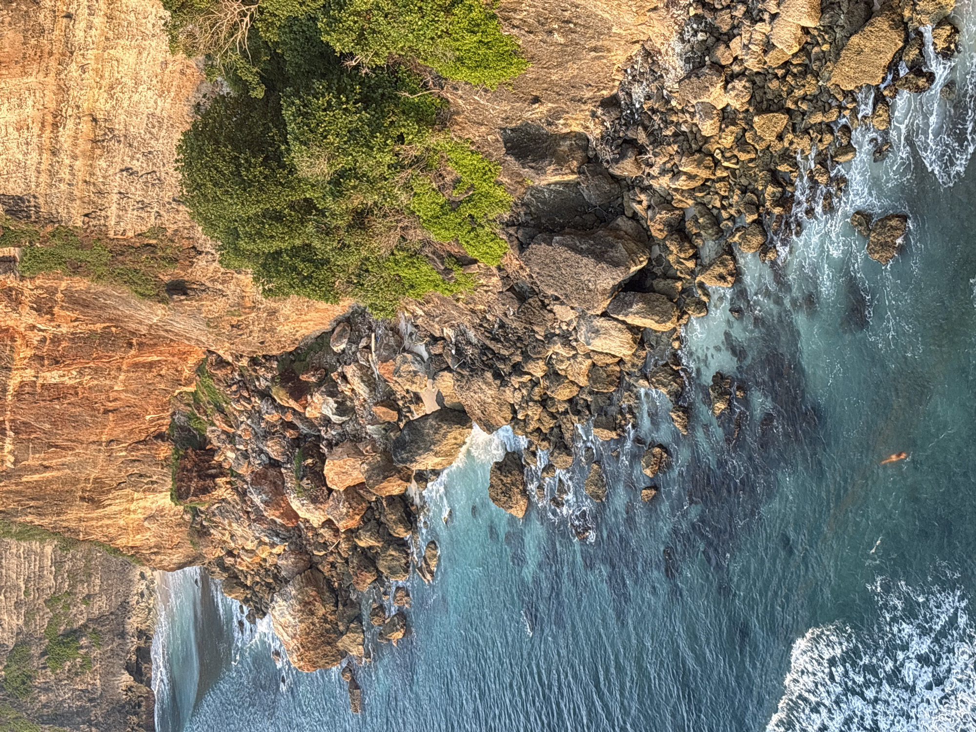 An aerial view of a rocky coastline with a high cliff in Pecatu, Indonesia, bordering the ocean. The cliff face on the left side of the image is a mix of brown and orange tones and covered with green vegetation on the top. Below the cliff, there's a rocky area with many large, brown-colored rocks. The water near the rocks is light blue and clear, becoming deeper blue further away from the shore. White foamy waves crash against the rocks, and a person is swimming in the water.