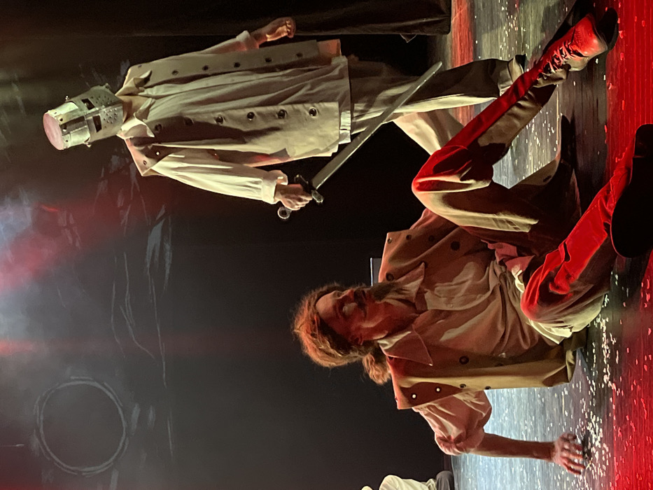 The scene depicts a theatrical performance unfolding on a stage in Novoshakhtinsk, Russia. Two male figures are central to the composition. In the foreground, one figure is seated on the dark stage floor, leaning backward with his left hand supporting him and his legs extended forward. He wears a light-colored long-sleeved shirt, a tan sleeveless vest with dark circular details, dark trousers, and dark lace-up boots. His hair is reddish-brown, tied back, and he sports a prominent mustache and goatee. His head is turned to his right, looking upwards, with an intense expression. Behind and to the right of the seated figure, a second individual stands, facing generally towards the foreground figure. This standing figure wears a light-colored long-sleeved shirt and a tan sleeveless coat-like garment. His head is fully covered by a metal bucket-style helmet, featuring horizontal eye slits. He holds a silver-colored sword with a dark hilt, pointed downwards, in his right hand. The dark stage floor is speckled with small white debris, resembling confetti. The background is a dark theatrical backdrop adorned with abstract white brushstroke-like markings, including a distinct large white circle in the upper left portion. The scene is illuminated by dramatic stage lighting, with notable red and white spotlights.