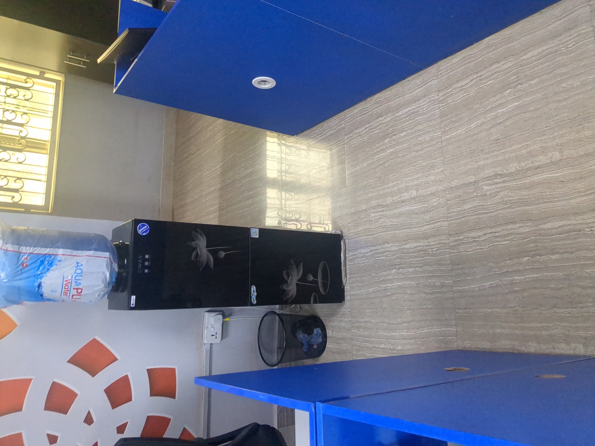 The image depicts a water cooler with a blue water jug on top, situated in what appears to be an office or communal space. The water cooler is black with decorative white lotus flower motifs. Next to the water cooler is a black mesh trash can containing some blue and black debris. A white electrical outlet with a yellow cord plugged into it is visible on the wall behind the trash can. The floor is tiled with a light brown and gray striped pattern. To the left and right of the scene are blue desk dividers or cubicles, one of which has a circular white grommet. In the background, a window with decorative metal bars and a yellow tint can be seen. The overall impression is of a functional, possibly shared, workspace. The image does not provide any direct or indirect information about the city of Fagge A, Nigeria, or any specific location within it.