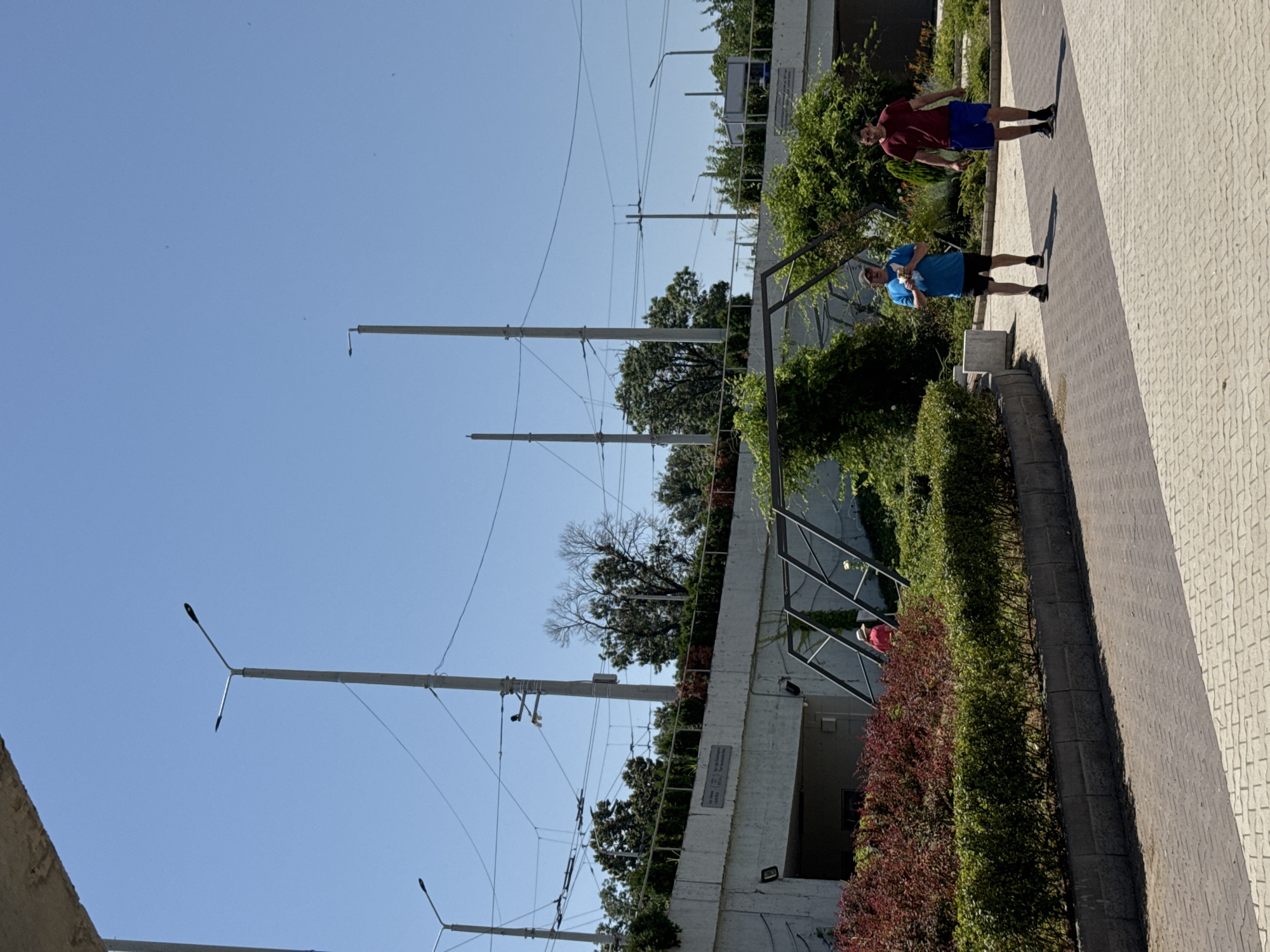 Two adult males are walking on a paved walkway in an outdoor urban setting. The male on the left is wearing a blue t-shirt and black shorts, and is holding a white object in his right hand. The male on the right is wearing a burgundy t-shirt and blue shorts. Both individuals are facing forward and appear to be walking in the same direction. Behind them, there is an elevated concrete structure with a green hedge and red bushes planted along its edge. Overhead, there are power lines and lampposts. The sky is clear and blue. This scene is located on the western part of the Ruse city center, near the Danubian Park, with the presence of the old tram infrastructure which is still partially visible in the background.