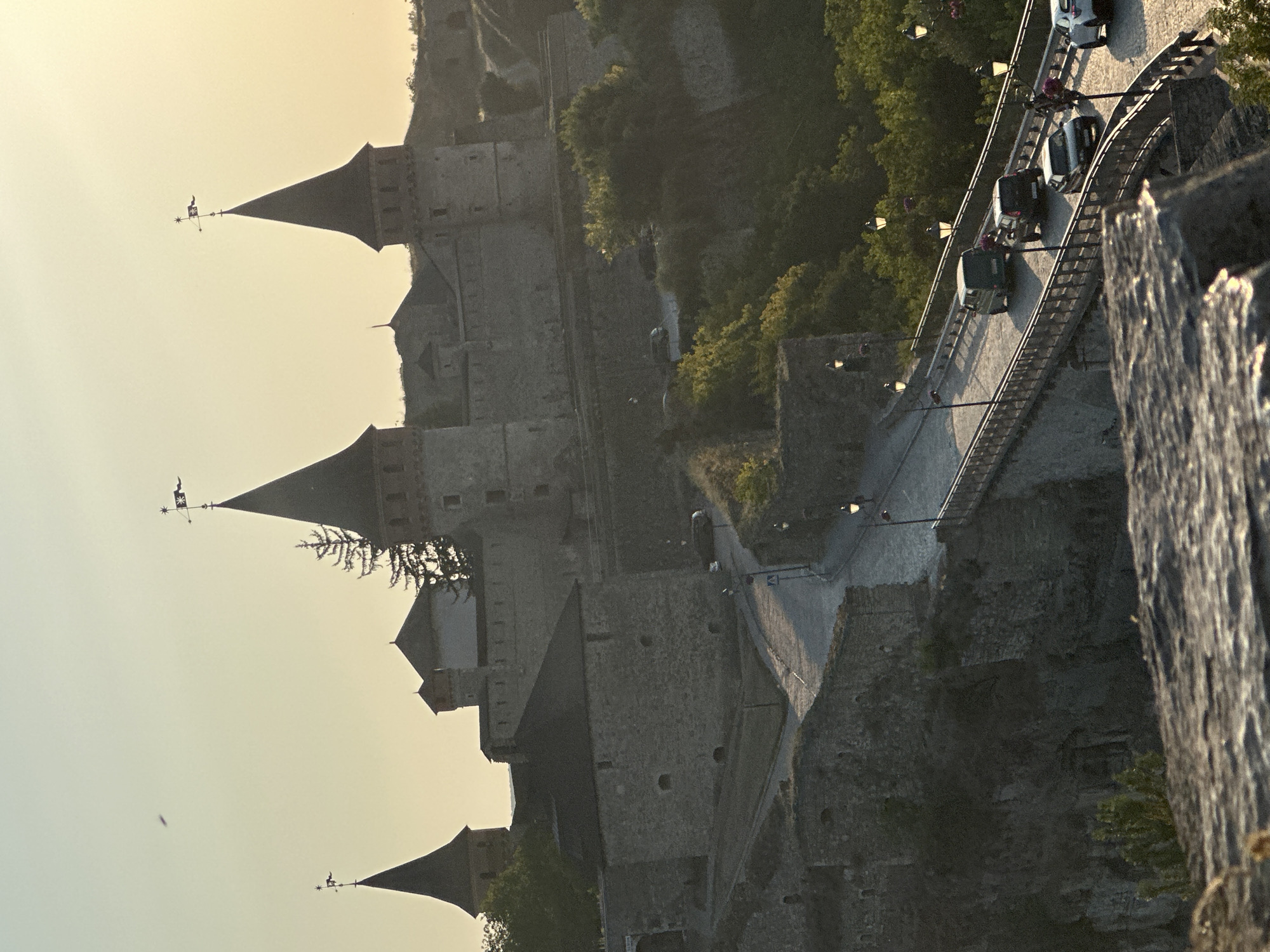 This image captures a view of the Kamianets-Podilskyi Castle in Ukraine, likely during the golden hour due to the warm, soft lighting and long shadows. The castle, a prominent historical landmark, is characterized by its stone construction, numerous towers with conical roofs, and battlements. In the foreground, a stone bridge arches over a lower level, with several cars driving across it. The bridge is lined with lampposts, some of which are illuminated. The surrounding landscape features lush green trees and rocky terrain. The sky is a pale yellow, indicating either sunrise or sunset. The overall scene depicts a blend of historical architecture and modern life, with the castle serving as a majestic backdrop to the ongoing activity on the bridge.