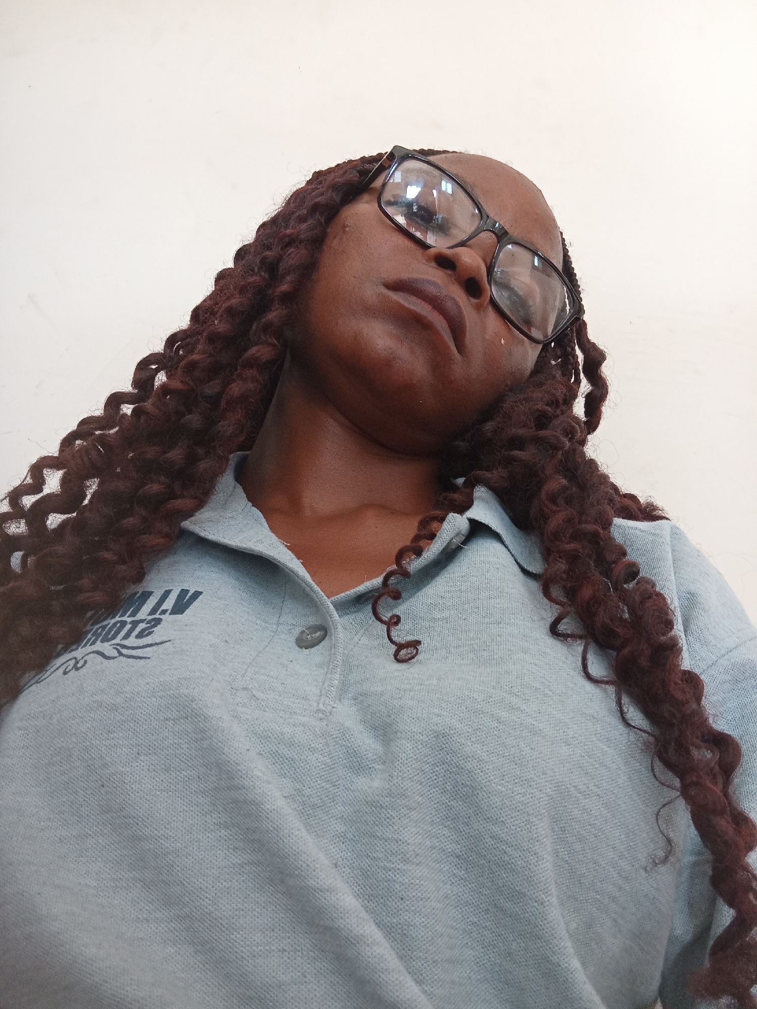 A low-angle, close-up shot shows a person wearing glasses and a light blue polo shirt. The person has dark skin and long, dark, curly braided hair. The glasses have black frames. The polo shirt has a collar and a button closure, with dark text printed on the left side. The text appears to be in a stylized font and is partially obscured by the person's hair and the angle of the shot, but some readable elements include "L.V." and "ROTS". The background is a plain, light-colored wall. The lighting suggests it is daytime, and the setting appears to be indoors. The person's expression is neutral or slightly contemplative.