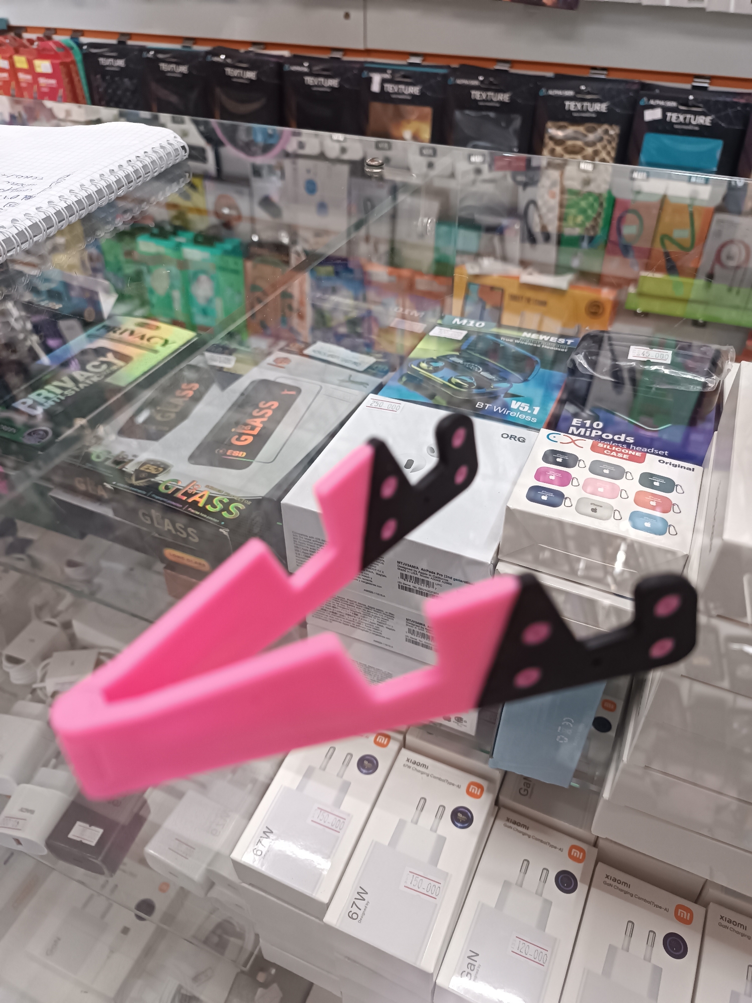 The image depicts a retail display case filled with various electronic accessories in a shop located in Chirchik, Uzbekistan. Prominently featured in the foreground is a bright pink phone stand. Behind the stand, the display case contains several boxes of screen protectors labeled "GLASS" and "PRIVACY," as well as packaging for wireless earbuds and phone chargers, many bearing the "Xiaomi" brand. The items are arranged on glass shelves, and the background shows rows of other packaged accessories.