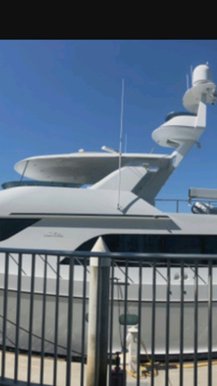 A blurry, wide shot captures the top section of a white yacht against a clear blue sky. The yacht is positioned horizontally, with its sleek, modern design featuring multiple levels and antennas. A railing made of dark metal bars runs across the foreground, obscuring the lower portion of the yacht. The background is uniformly blue, indicating an outdoor setting with no discernible landmarks or cityscape visible, thus rendering the location within Hongkou District, China, unidentifiable from the visual information provided. No people or specific actions are depicted.