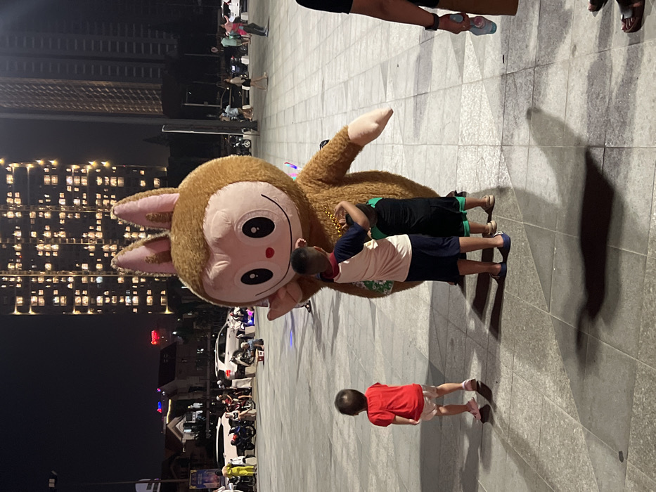 This nighttime image captures a lively urban public square, identified as the plaza at Vinhomes Central Park, adjacent to Landmark 81, in the Binh Thanh District of Ho Chi Minh City, Vietnam.
In the center, a person in a large, brown, furry mascot costume, resembling a monkey or rabbit with a light pink face, large dark eyes, and a wide smile, stands with its left hand raised in a waving gesture. The mascot wears gold-colored bead necklaces. Two young children, seen from behind, are embracing the mascot at its midsection. The child on the left wears a light-colored shirt with dark sleeves and dark shorts; the child on the right wears a dark shirt and shorts. To their left, a third young child in a red shirt and light shorts stands with their back to the camera, facing the mascot. Part of an adult holding a light blue bottle is visible on the far right foreground.
The ground consists of large, light gray square and rectangular paving tiles, showing multiple elongated shadows from the subjects and ambient lighting.
The background on the left features several parked motorcycles and cars, with groups of people gathered around. Beyond them, a row of low-rise buildings with illuminated windows is visible, along with a partial red neon sign on a distant building. The background on the right is dominated by a very tall, dark skyscraper, clearly identifiable as Landmark 81, with numerous brightly lit windows across its facade, indicating residential or office occupancy. An adjacent, less illuminated high-rise is also present. The scene is artificially lit, creating distinct light and shadow patterns.