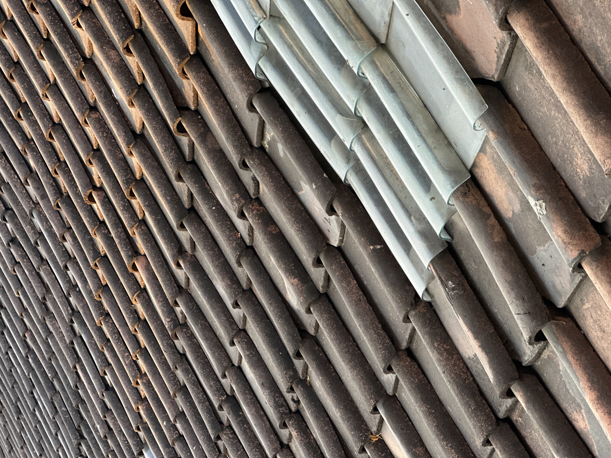 This is a close-up, aerial photograph of a tiled roof in Bulakrejo, Indonesia. The roof is composed of two distinct sections. The larger section on the left is covered with weathered, dark brown terracotta tiles laid in overlapping rows. These tiles show signs of age, with some dust and debris accumulated on their surfaces. To the right, there is a section of corrugated, translucent roofing material. This material is light gray and appears to be made of plastic or fiberglass, with a wavy pattern. The two types of roofing materials meet at an angle, creating a clear demarcation line across the image. The sunlight casts shadows, highlighting the texture and contours of the tiles and the corrugated material. The background is not visible, focusing entirely on the rooftop's composition.