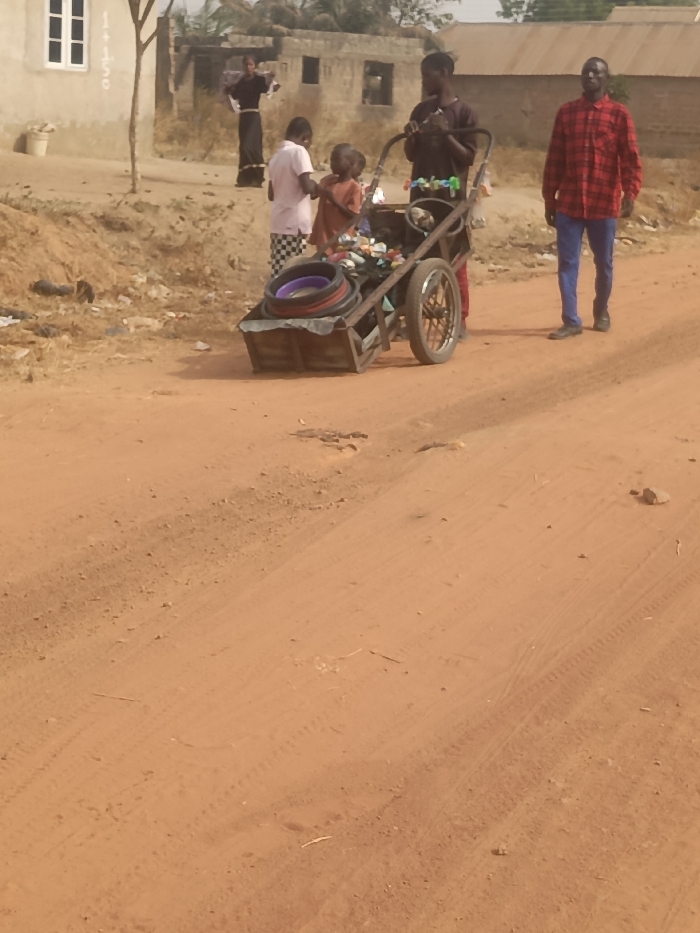 A sandy path leads to a group of people. One man wears a red plaid shirt and blue pants, walking toward the camera. Two children are with a man pushing a cart filled with assorted items, including tires and plastic containers. Another person is standing in the background, dressed in dark clothing. Buildings are visible in the background. The scene is sunlit, creating shadows on the sandy ground.