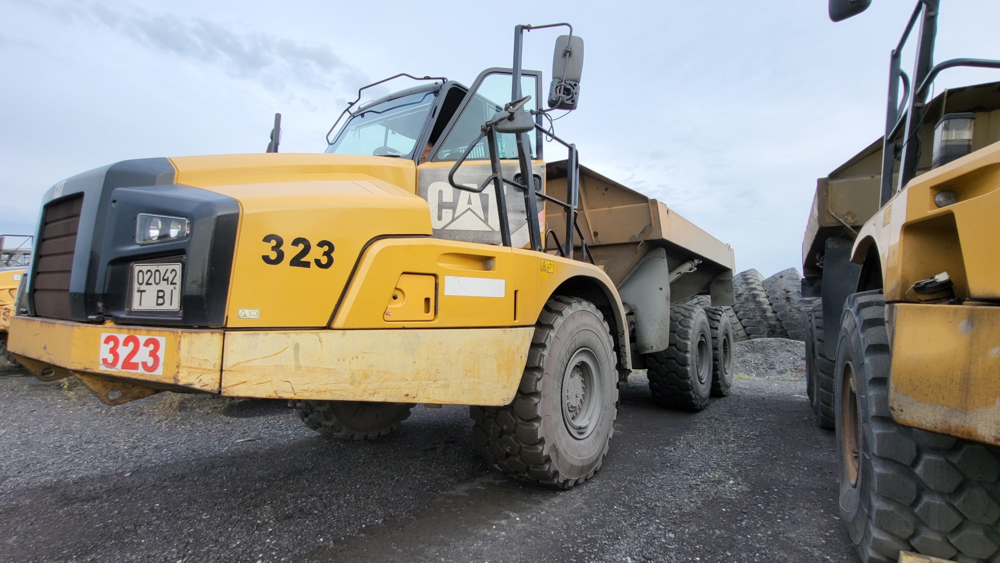 This image shows a close-up view of two yellow articulated dump trucks parked on a gravel surface, likely at a construction or industrial site. The truck in the foreground is a Caterpillar model, identified by the "CAT" logo on its side and the number "323" prominently displayed in black on the yellow body and in red on the front bumper. This truck has large, rugged tires. A license plate with the numbers "02042" and letters "T BI" is visible on the front of the truck. The second dump truck is partially visible to the right, also yellow, and appears to be of a similar make and model. In the background, a pile of large tires is visible, possibly spare tires for the heavy machinery. The sky is overcast. The image is undated and provides no specific information about the city of Voloshyne, Ukraine, beyond its inclusion in the prompt.