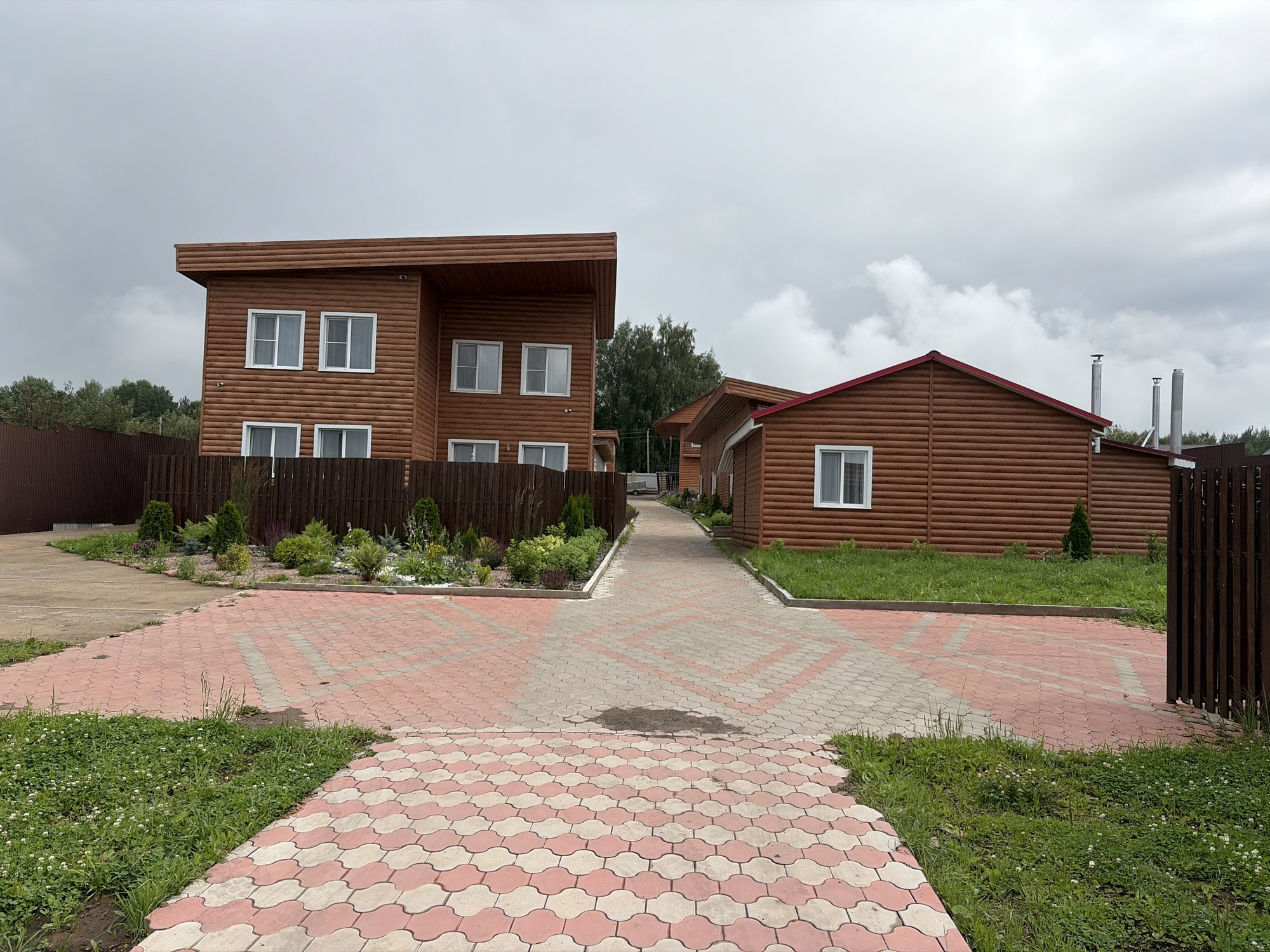 The image shows a street with two wooden houses on a cloudy day. The houses are modern with large windows and dark brown wood siding. The street is paved with pink and white interlocking pavers, and there is grass on either side of the street. The houses are surrounded by a wooden fence. The location is the city of Пакли, Russia.