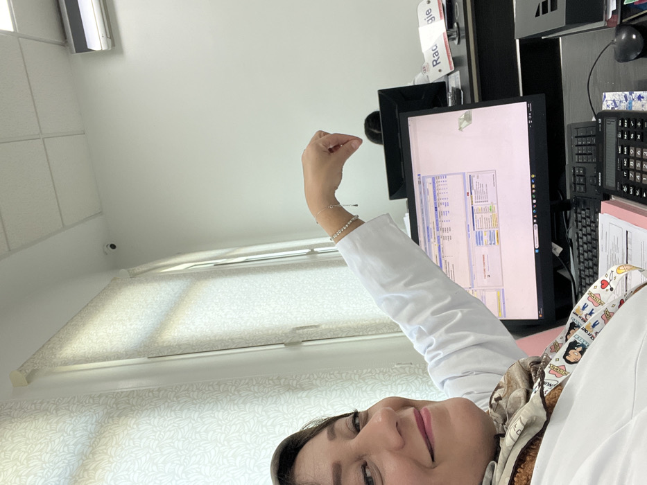 A woman wearing a white coat is posing for a selfie in what appears to be a medical office. Behind her is a computer monitor displaying lines of data. To the monitor's right is a calculator and a keyboard. The background includes a window with white blinds. A security camera is visible on the ceiling. The woman is wearing bracelets and has her hand raised near the monitor.