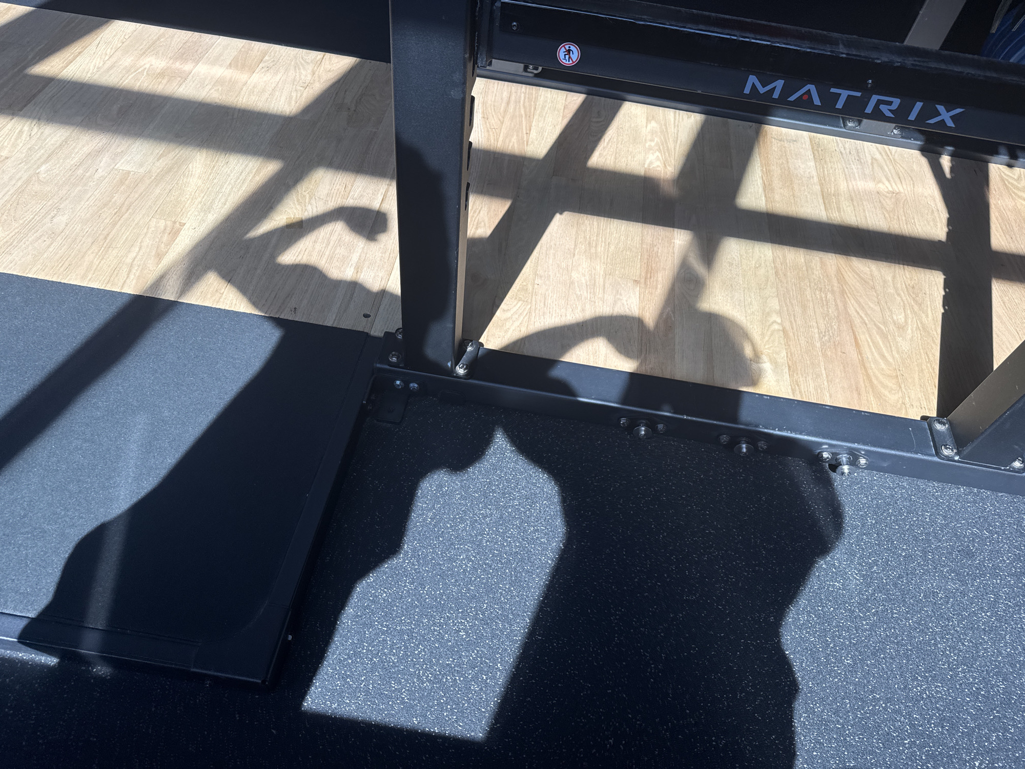 The image shows a shadow of a flexed bicep on a wooden floor, cast by sunlight filtering through a gym rack. The gym rack has the brand name "MATRIX" visible in white lettering. The floor is made of light-colored wood planks, with a black rubber matting area in the foreground. A black metal frame is part of the gym equipment, with visible bolts. The shadow of the bicep is well-defined and appears to be that of a person's arm. The overall scene suggests a workout session in a gym.