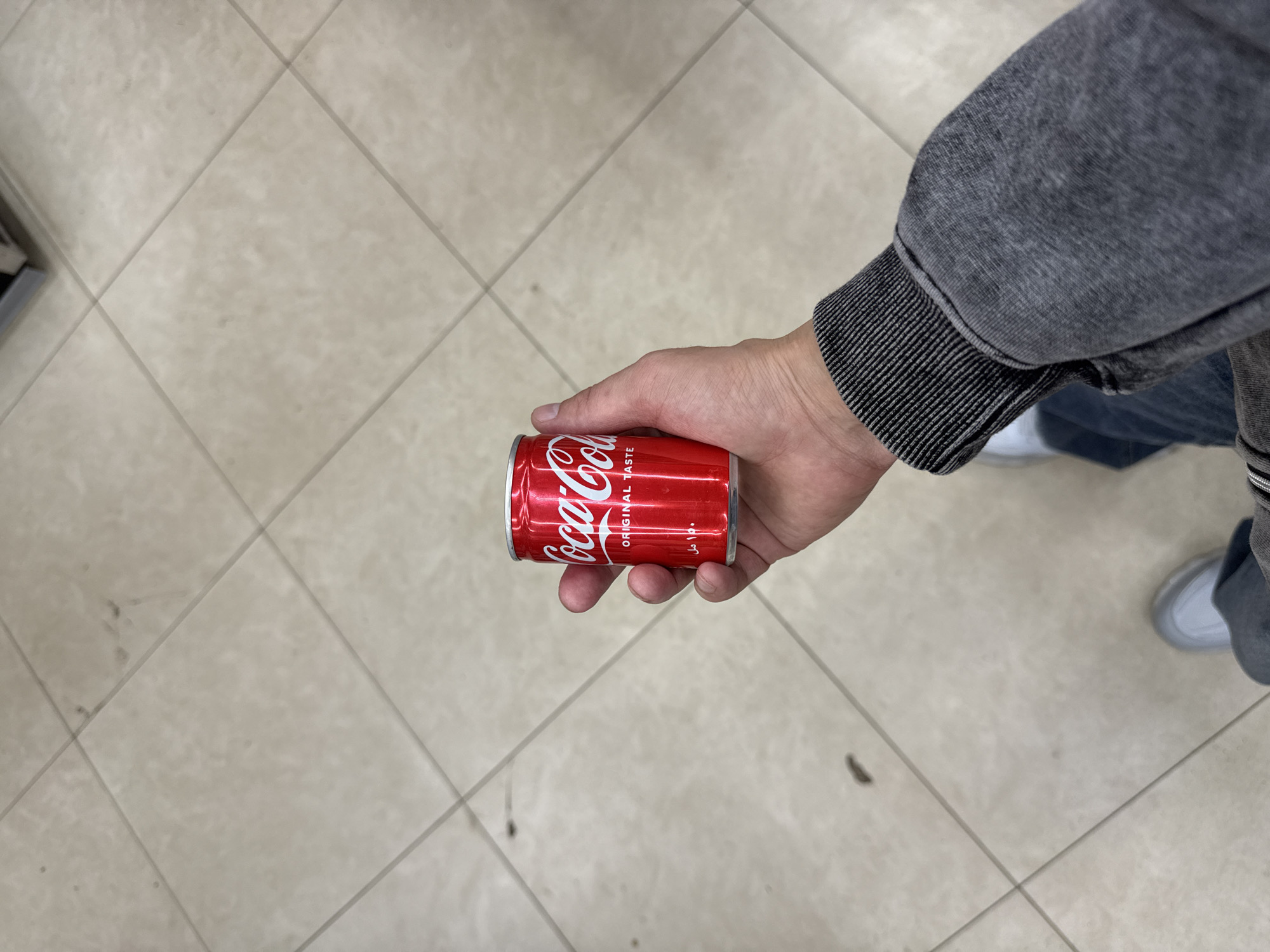 A person's hand is holding a red Coca-Cola can. The person is wearing a gray sweater and blue jeans. The background is a tiled floor. The location is not specified, and there is no information indicating it is Moscow, Russia.