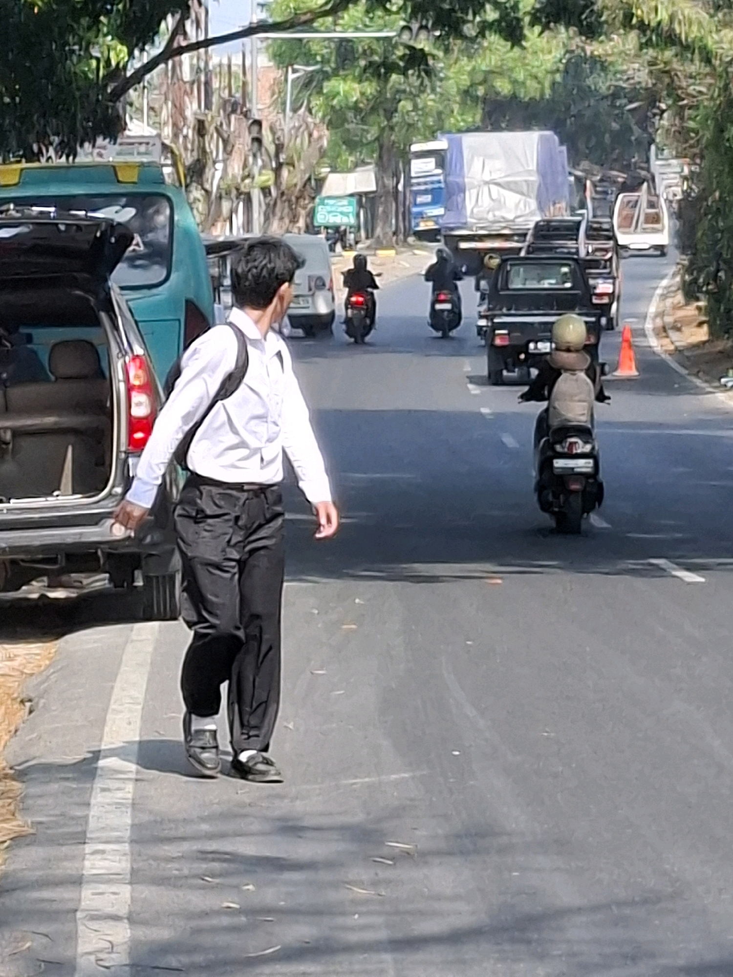 The image depicts a street scene in Sukabumi, Indonesia, with a man in a white dress shirt and black pants walking on the left side of the road. He is carrying a black backpack and holding a small object in his right hand. To his left, a teal-colored van with its trunk open is parked. The road is paved and has white lane markings. Several vehicles are visible in the background, including a black pickup truck, multiple motorcycles, a large truck covered with a blue tarp, and a white vehicle. Green trees line the right side of the road, and buildings and utility poles are present further in the background. The lighting suggests it is daytime.