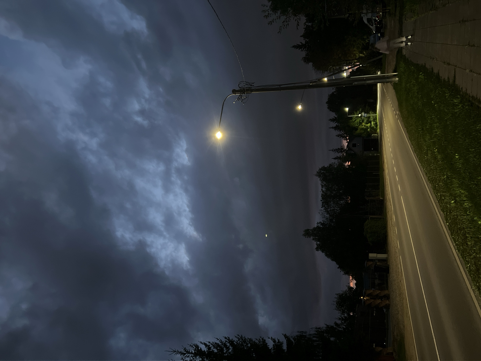 A view of a street in Vilnius, Lithuania, at dusk with dark, stormy clouds overhead. A paved road runs through the scene, with a sidewalk on the right side. A person is rollerblading on the sidewalk, facing away from the camera. Streetlights illuminate the road and sidewalk. Trees and houses are visible in the background.