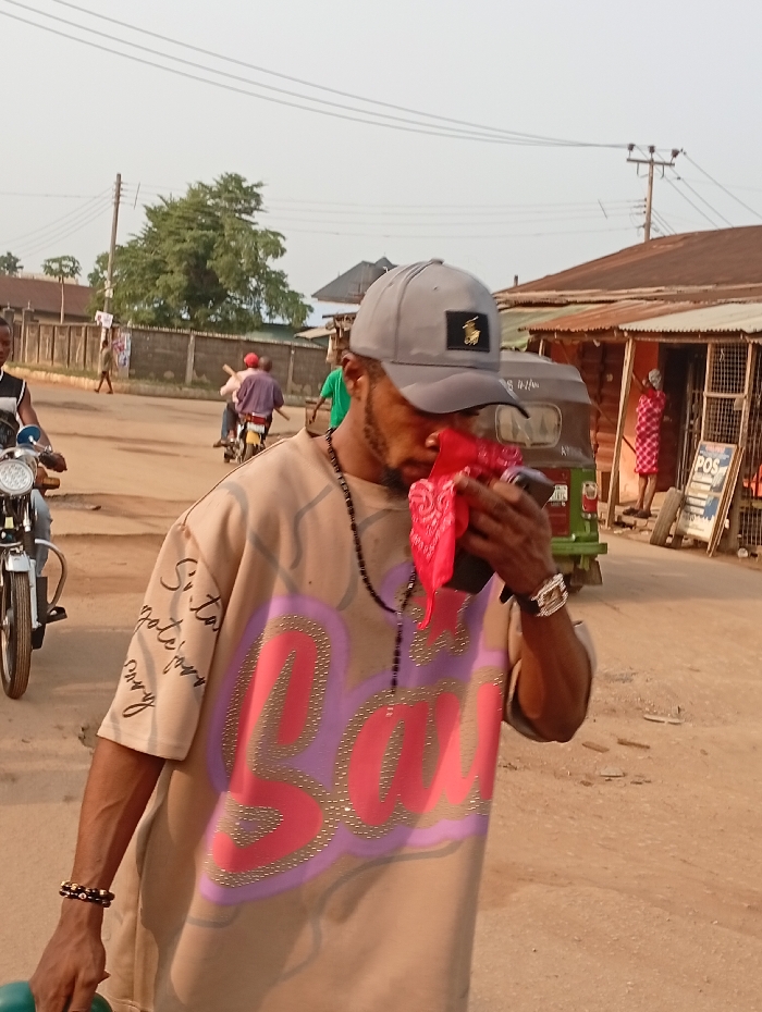 This image captures a street scene in Umuode, Nigeria, under daylight conditions. In the foreground, a bearded male wears a grey baseball cap with a dark and gold emblem, a black beaded necklace, and a loose-fitting, beige t-shirt adorned with pink and light purple rhinestone text (partially reading 'Sauce' or 'Baddie'). Script is visible on his left sleeve. He holds a red bandana and a dark object in his right hand, partially covering his lower face, and a green object in his left hand. A silver-toned watch is on his right wrist, and a dark beaded bracelet on his left.
The unpaved street recedes into the background. To the left, a person on a motorcycle is partially visible, followed by two individuals riding another motorcycle, one carrying a long stick. A green and black Keke Napep (tricycle taxi) is parked on the right side of the street, in front of a row of single-story shops or market stalls with corrugated metal roofs. One shop displays a "POS" sign. A person in a red and white patterned dress stands outside one of these shops. Utility poles with overhead lines and several trees are visible in the background, set against a light sky.