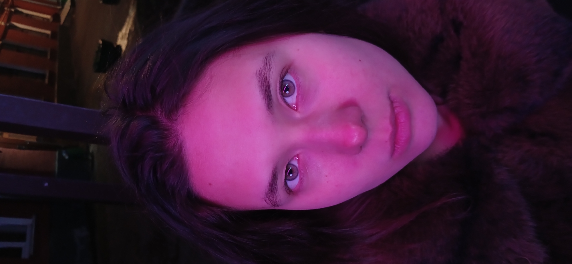 A young person, likely female, with dark hair and light-colored eyes, is depicted in a close-up portrait, rotated 90 degrees counter-clockwise. Her face is prominently lit by a strong pink and purple light, casting a vibrant glow on her skin and hair, while the surrounding environment remains largely dark and out of focus. She looks directly into the camera with a neutral and somewhat pensive expression. Her lips are slightly parted, showing a hint of a smile or a relaxed pose.
Her dark hair frames her face, with strands visible against the dark background. She appears to be wearing a dark, possibly furry, garment or jacket, visible on the right side of the frame. The background is indistinct but shows blurred shapes that suggest an indoor setting, possibly with some wooden elements or furniture in the upper left, such as what might be a musical instrument (xylophone) or benches. The overall lighting scheme creates a moody and artistic atmosphere. No specific activity is occurring beyond posing for the photograph. There is no visible text. The image likely depicts a scene in Moscow, Russia.