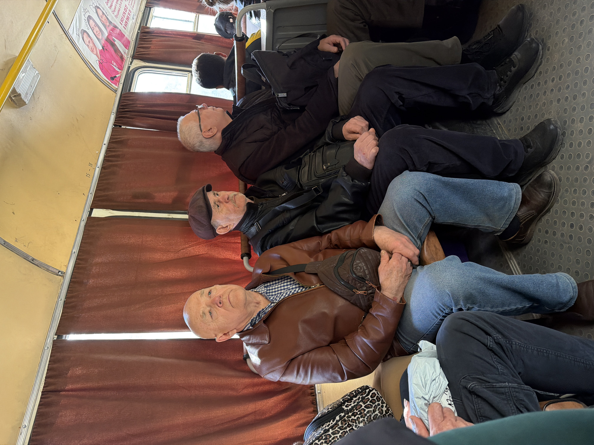 The image shows a scene inside a public transport vehicle, likely a bus or a tram, in Noviy, Russia. Four older men are seated next to each other.
The closest man to the camera has a bald head and wears a brown leather jacket over a checkered shirt, jeans, and brown leather shoes. He also has a small dark leather bag on a strap across his chest. Next to him is a man wearing a dark leather jacket, a brown cap, blue jeans, and brown boots. The third man wears glasses and a dark jacket. The fourth man has a black backpack.
The seating is upholstered with a brown fabric. Above the seats, there are advertisements on the walls. The floor has a textured gray surface. The lighting suggests it is daytime.
