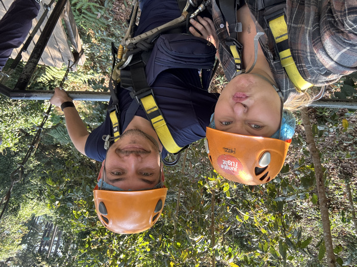 Two individuals, one female and one male, are depicted on an elevated platform in a dense jungle environment within the city of Pa Miang, Thailand, consistent with a zipline or canopy adventure activity. Both wear orange Petzl climbing helmets over blue hairnets and yellow full-body safety harnesses. The female, positioned closer to the camera on the left, has light-colored hair and is wearing a plaid shirt in shades of brown, grey, and white. She is making a puckered, pouting facial expression directly at the camera. The male, positioned to the right, has dark hair and stubble, and is wearing a dark blue short-sleeved shirt. He is smiling towards the camera. Both individuals are partially grasping a silver metal railing in the foreground. Behind them, lush green foliage dominates the background, with further wooden and metal platform structures visible to the right, descending into the forest. The female's helmet features a "Jungle Flight" sticker.