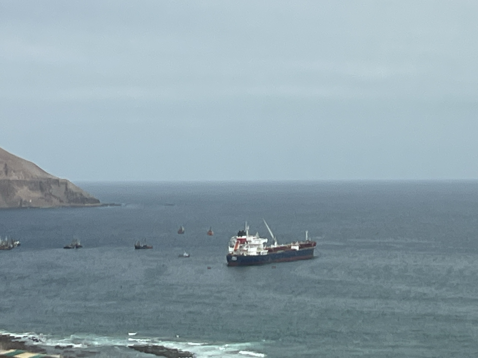 The image shows a wide shot of the ocean off the coast of Arica, Chile. Several ships are visible, including a large cargo ship in the center of the frame. The cargo ship is dark blue and red with a white superstructure and a crane. Smaller ships, some appearing to be fishing boats or tugboats, are scattered in the water. In the background, a rocky, barren coastline is visible, characteristic of the coastal desert landscape of the region. The sky is overcast and a pale blue. The water is a deep blue-green and appears somewhat choppy. In the foreground, a blurry view of waves crashing on what appears to be a breakwater or rocky shoreline is visible. The overall impression is a scene of maritime activity in a rugged coastal environment.