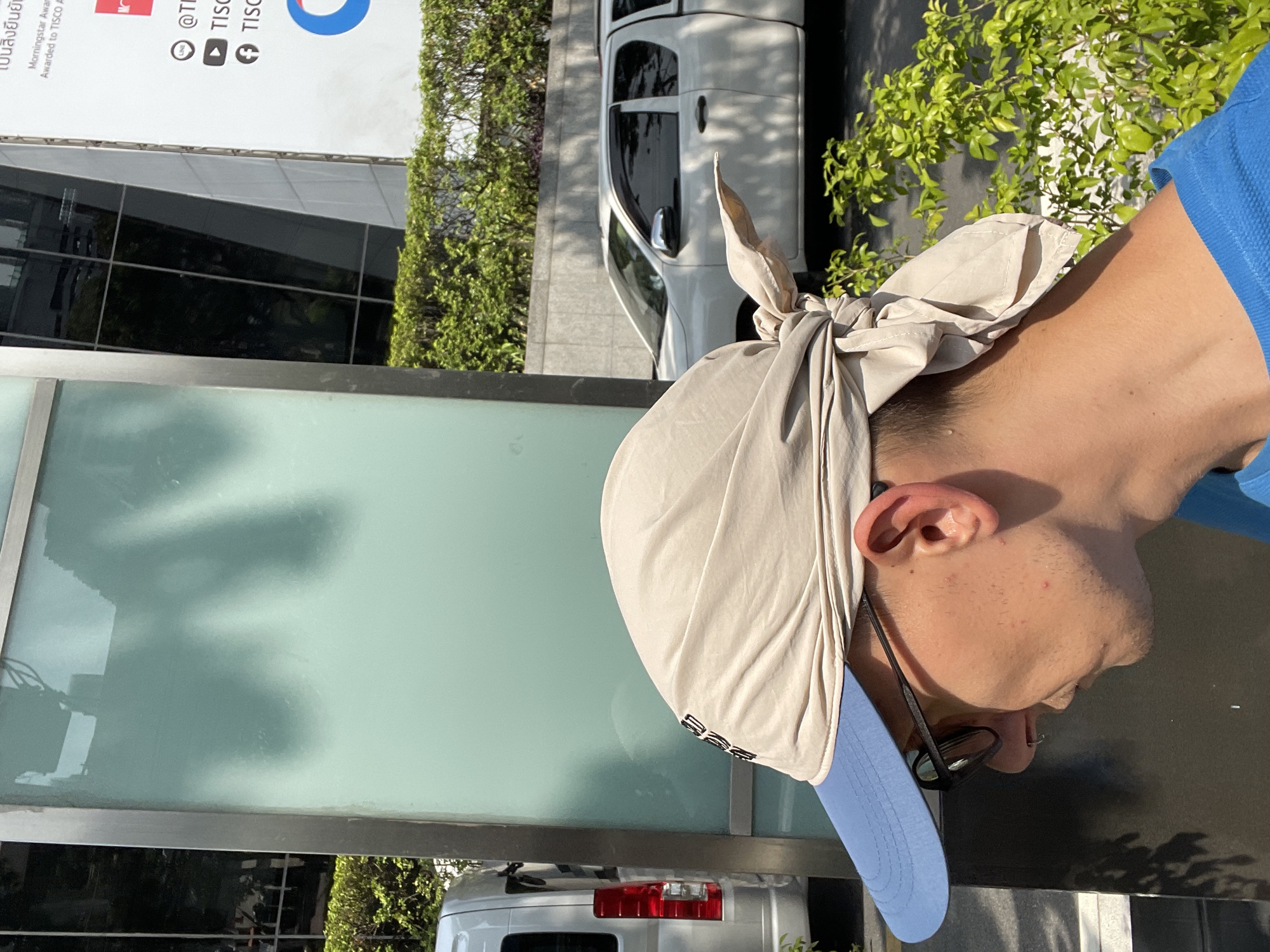 A street-level, sunlit view focuses on a person wearing a blue polo shirt, glasses, and a light beige head covering tied at the back of the head with a long blue brim. The person is standing in front of a large glass window that reflects a street scene. Behind the person and to the left of the window, parts of two white vans or large vehicles can be seen. Lush green foliage is present on both sides of the frame, providing a natural backdrop. A building with text in Thai and English is visible in the upper left of the frame with the text "Morningstar Award". The location is likely in Bangkok, Thailand, given the presence of Thai language.
