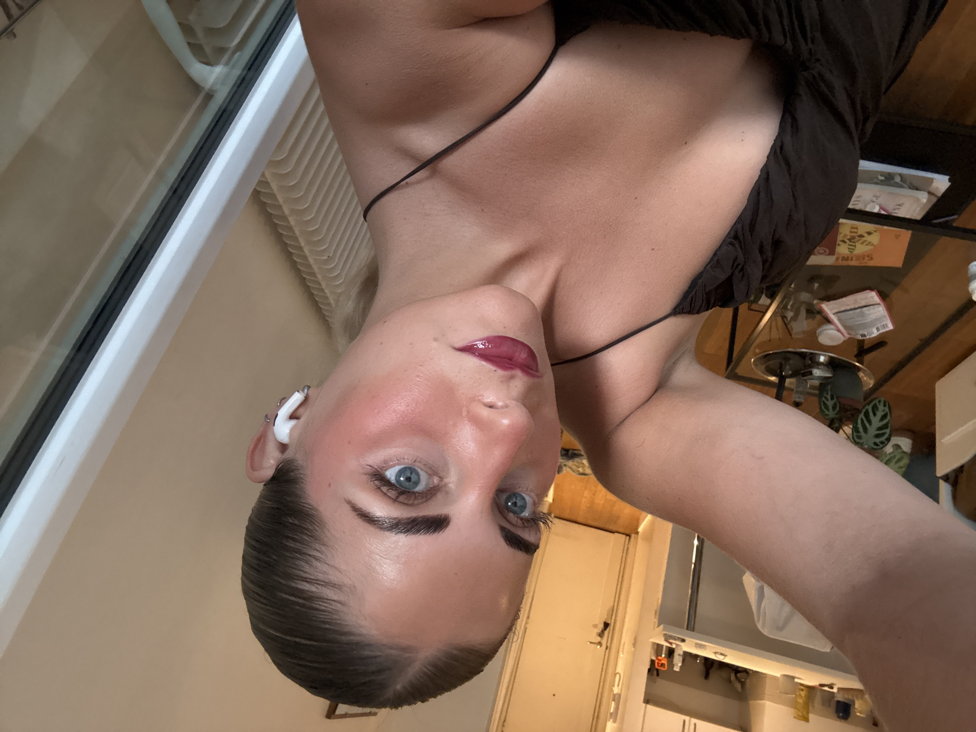 A young woman with fair skin, blue eyes, and dark hair pulled back is taking a selfie. She is wearing a dark brown spaghetti strap dress and white wireless earbuds. Her makeup includes a glossy berry-colored lipstick and noticeable pink blush on her cheeks and nose. She has styled eyebrows and long eyelashes. In the background, a cluttered table with various items, including a plant, is visible. A doorway and part of a kitchen area with cabinets are also present. The scene appears to be indoors, possibly an apartment. The location is not Paris, France.