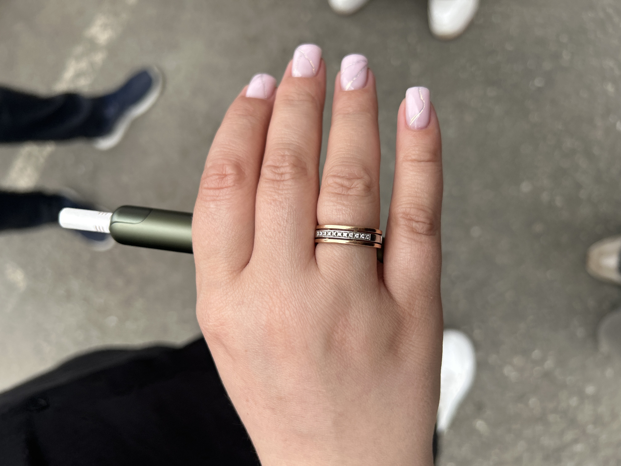 The image shows a close-up of a person's hand, adorned with a rose gold ring featuring inlaid diamonds, and manicured nails painted a light pink with a marble-like design. The hand is holding a green and white electronic device. In the blurred background, two individuals are visible wearing dark pants and athletic shoes. The ground appears to be asphalt or concrete, suggesting an outdoor urban setting, possibly in the city of Perm, Russia.
