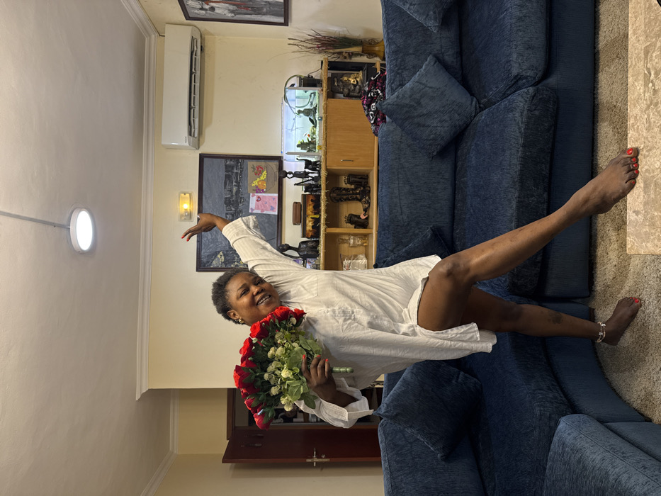 The image depicts an indoor scene, likely a living room, in Alaguntan, Nigeria. An adult female, appearing to be of African descent with short dark hair, stands prominently in the center. She wears an oversized white collared shirt, functioning as a dress, and has red nail polish on her fingers and toes. A silver-colored ankle bracelet is visible on her left ankle. She is smiling and looking towards the camera, with her left leg bent and her right leg extended forward, foot flat on the ground. Her left arm holds a large bouquet of red roses with green foliage and white filler flowers, while her right arm is raised and bent at the elbow.
The background features a dark blue sectional sofa with matching throw pillows on the left and mid-ground. The floor is covered with a light brown carpet, and a light-colored rectangular coffee table is partially visible in the foreground. The walls are cream-colored with white crown molding. On the left, a dark wooden door is partially ajar. Along the back wall, a wooden console table or cabinet is adorned with various objects: several dark figurines depicting human and animal forms (including a giraffe), a small fish tank containing fish, a brown box, some white items, a pair of dark boots, and a statue resembling a meditating figure. To the far right, dried decorative branches are in a vase. Above the cabinet, a large dark-framed painting of a cityscape is centrally mounted, flanked by a yellow emergency exit sign above it and two smaller colorful cards below it on the wall. To the right of the painting, a white wall-mounted air conditioning unit is installed, and further right, another dark-framed abstract artwork with red accents is visible. The white ceiling features a single circular flush-mount LED light fixture.
