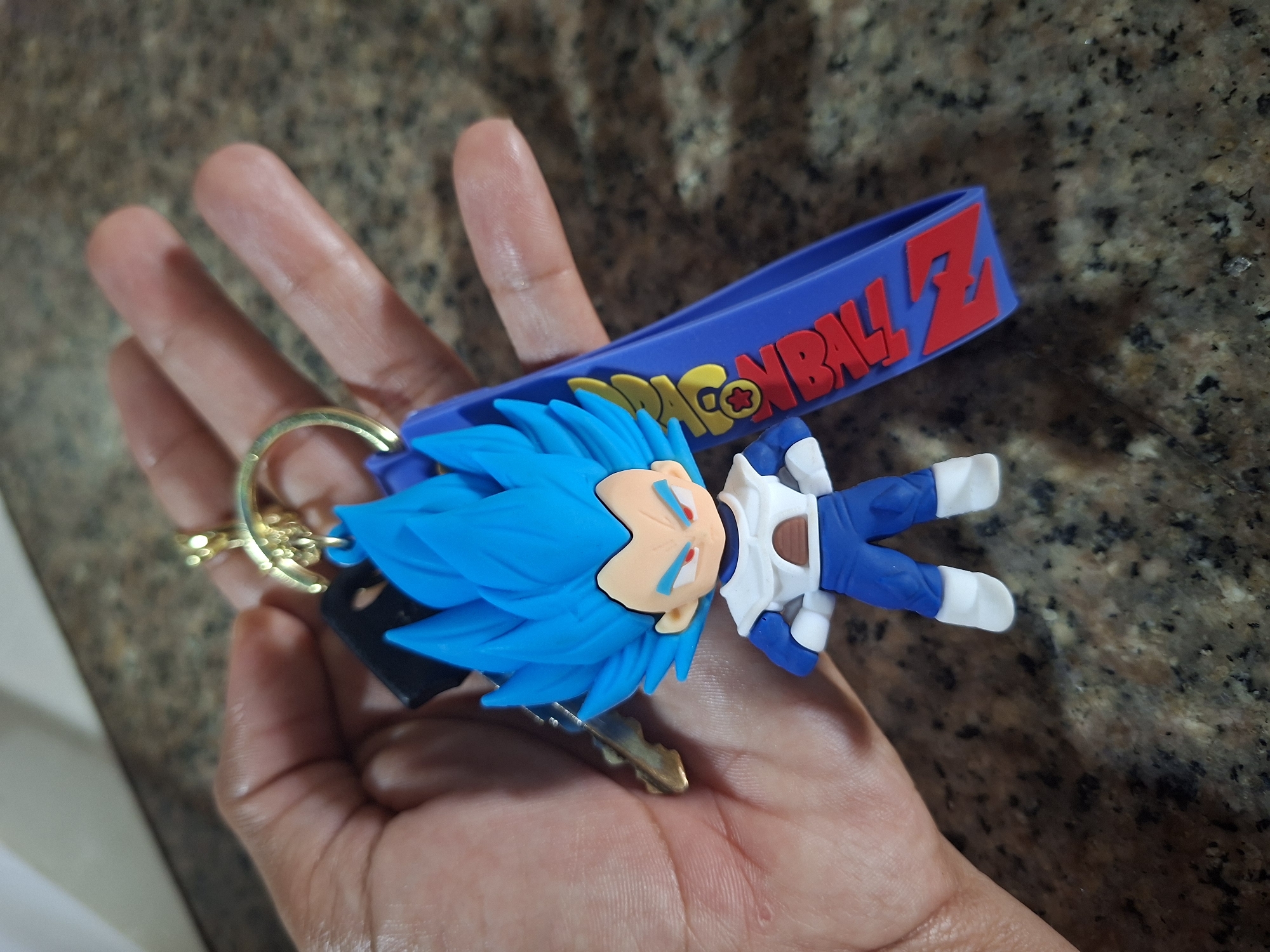 A close-up shot shows a hand holding a keychain with a Dragon Ball Z character. The character is a miniature figurine of Vegeta, a popular character from the anime series, with bright blue spiky hair and an orange and white outfit. The figurine is attached to a blue lanyard that features the "Dragon Ball Z" logo in red and yellow lettering. The background is a speckled granite countertop, suggesting an indoor setting. The lighting is bright, casting subtle shadows. There is no indication of the city of El Valle del Espíritu Santo, Venezuela in the image or its context.