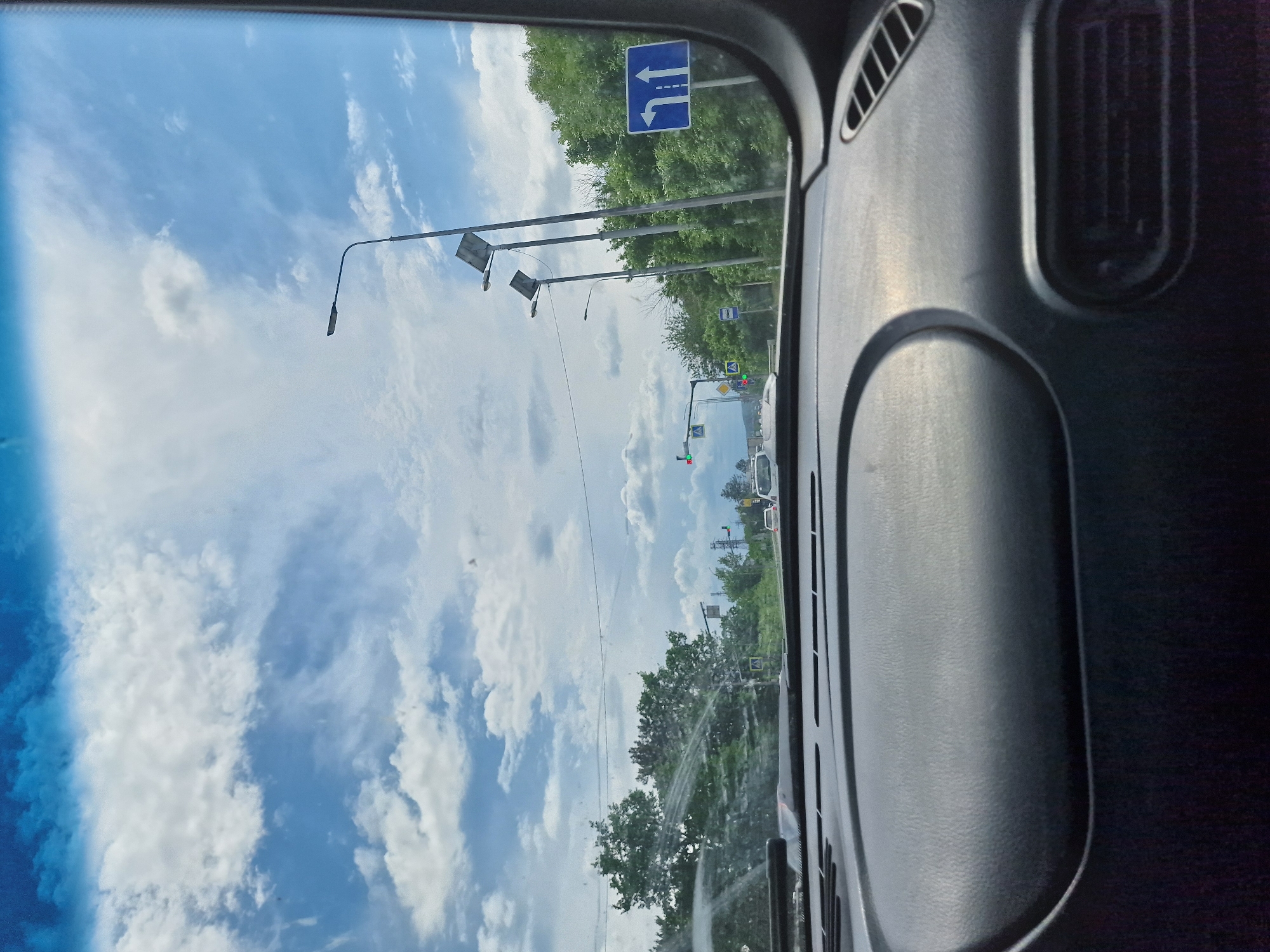 The image shows a view from inside a vehicle looking through the windshield at a road in Zmeyka, Russia. The sky is blue with scattered white clouds. There are trees lining the road, and several lampposts are visible. Traffic lights and road signs are also present. The dashboard of the car is in the foreground.