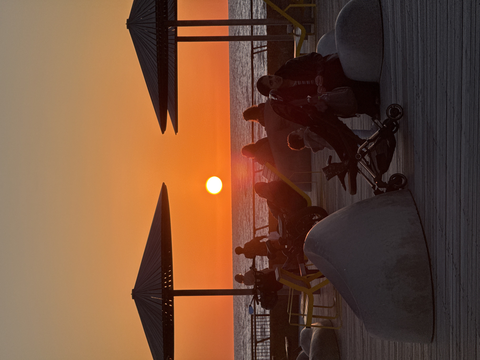 The image captures a vibrant sunset over a body of water, likely the Mediterranean Sea, from a modern boardwalk or pier in what is consistent with Tel-Aviv, Israel. The scene is dominated by the dramatic colors of the sky, which transitions from a light beige on the left to a brilliant orange and deep gold closer to the horizon where the sun is setting.
A bright, circular, white-yellow sun is positioned low in the sky, casting long shadows and silhouetting the subjects. The water below the horizon appears dark and calm, reflecting some of the sky's light.
On the left side of the frame, two prominent, large, fan-like architectural structures, possibly sunshades or artistic installations, stand in dark silhouette against the colorful sky. These structures feature a slatted design. On the right side, a wide wooden boardwalk with distinct vertical planks extends towards the foreground.
Several groups of people are gathered on the boardwalk, most appearing as dark silhouettes against the bright backdrop. They are positioned to observe the sunset, with some seated on large, rounded, dark grey benches or concrete elements that are part of the boardwalk's design. Yellow metal railings or support structures are also visible along the walkway. A baby stroller is present near one group of individuals.
The overall atmosphere is peaceful and serene, conveying a moment of shared relaxation and appreciation for the natural beauty of the sunset. There is no visible text in the image. The time of day is clearly evening, during golden hour.