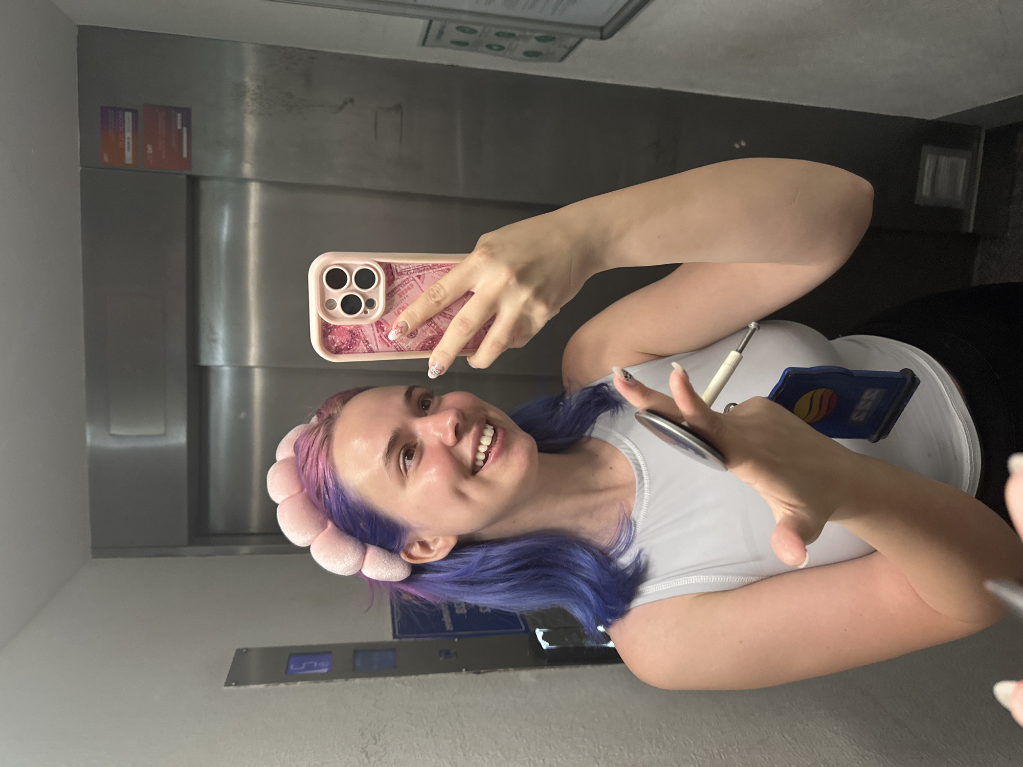 A woman with purple hair, wearing a pink headband and white tank top, is taking a selfie in an elevator. She is holding a pink iPhone in her right hand and a pen in her left. The elevator has stainless steel walls with elevator control buttons. The location is likely Ramonville-Saint-Agne, France, as the service card attached to her top might be a transportation service card.