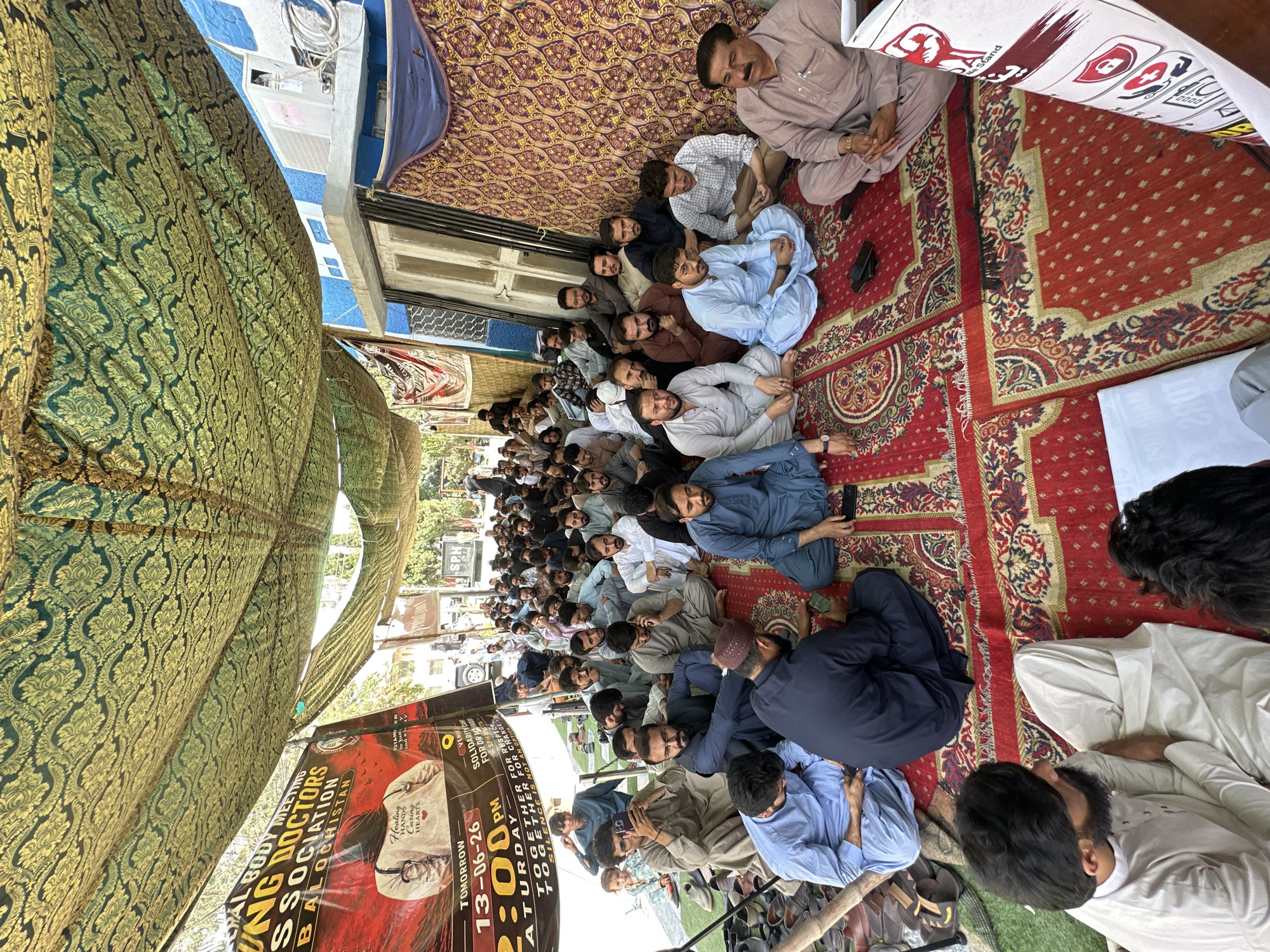 In Quetta City Tehsil, Pakistan, a gathering of individuals, primarily men, are seated on a red carpet under a patterned canopy. The scene appears to be an outdoor assembly. The individuals are dressed in traditional Pakistani attire, with many wearing shalwar kameez. Some are engaged in conversation, while others are looking at their phones or listening intently.
The canopy is made of a green and gold patterned fabric, creating a shaded area for the attendees. In the background, there are buildings, including one with a blue facade and an air conditioning unit. A banner with text and an image is visible on the left, indicating a "Young Doctors Association Balochistan" meeting scheduled for "Tomorrow 13-06-26" at "2:00 PM". Shoes are piled up on the left side of the image. The overall atmosphere suggests a community or professional gathering.