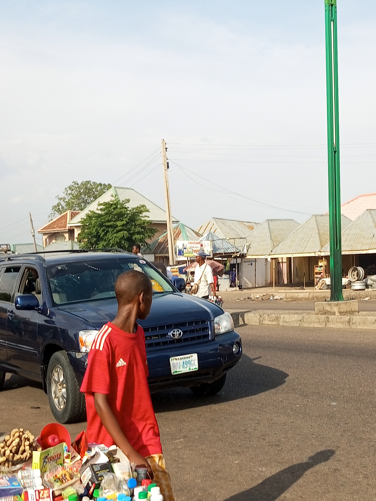 The image depicts a street scene in Gombe, Nigeria, featuring a dark blue SUV stopped on a paved road. A young boy wearing a red t-shirt with white stripes and an Adidas logo is positioned to the right of the vehicle, standing behind a collection of goods displayed on a table. These goods appear to be a variety of packaged items, including bottles and boxes, suggesting a small roadside stall. In the background, several market stalls with corrugated metal roofs are visible, along with a few people and a wooden utility pole. The sky is mostly clear with some light cloud cover. The overall environment suggests a busy urban or semi-urban market area.